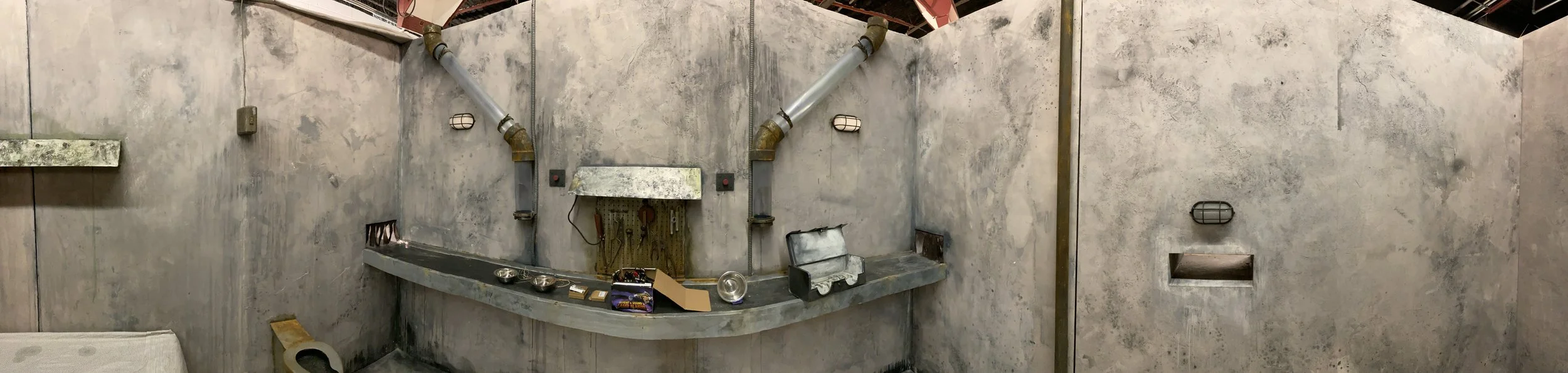 Film set of an industrial room with concrete walls, pipes, and various objects on a concrete countertop, including small tools, metal bowls, and a black case.