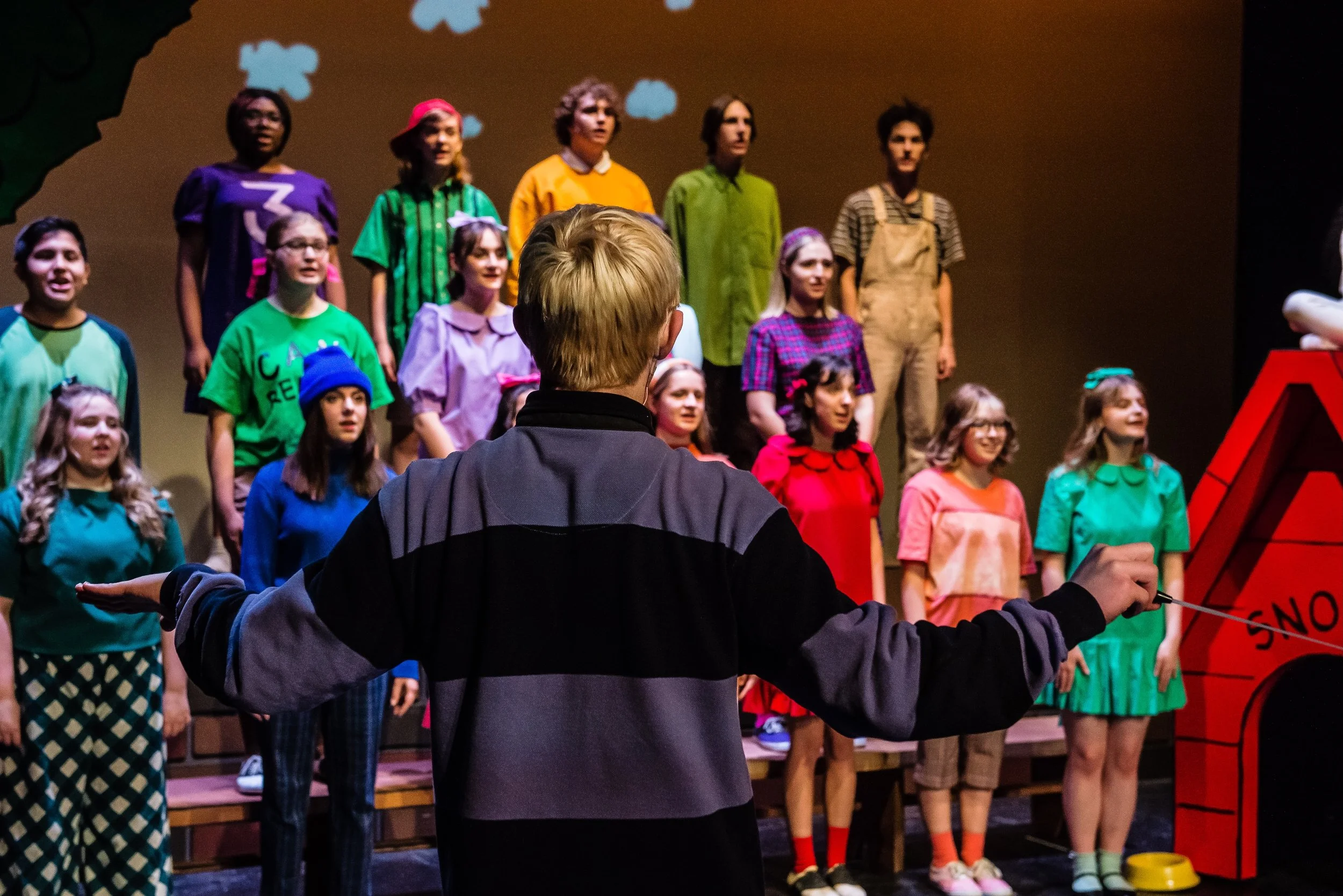 A performance of 'You're a Good Man Carlie Brown' with Schroder conducting a children's choir on stage, with kids dressed in colorful outfits singing.