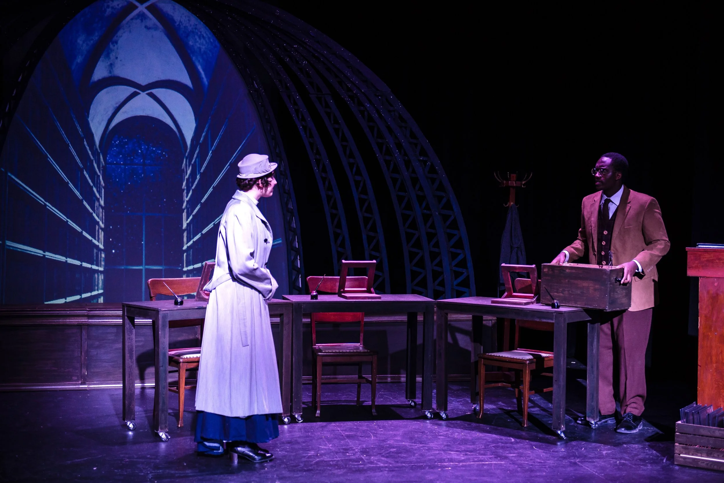 The image shows two actors on stage during a theater performance. The actor on the left is a woman dressed in a long beige coat and hat, facing the actor on the right. The actor on the right is a man wearing glasses and a brown suit, standing behind 