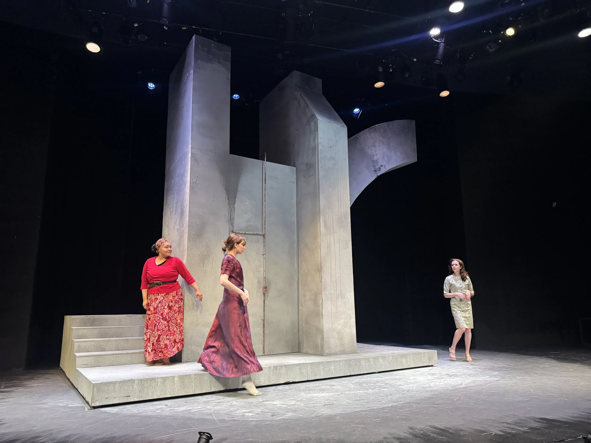 Three women are on a stage with a plain background and large concrete structures. They are part of a modern production of 'Romeo and Juliet', with one woman walking and the others standing.