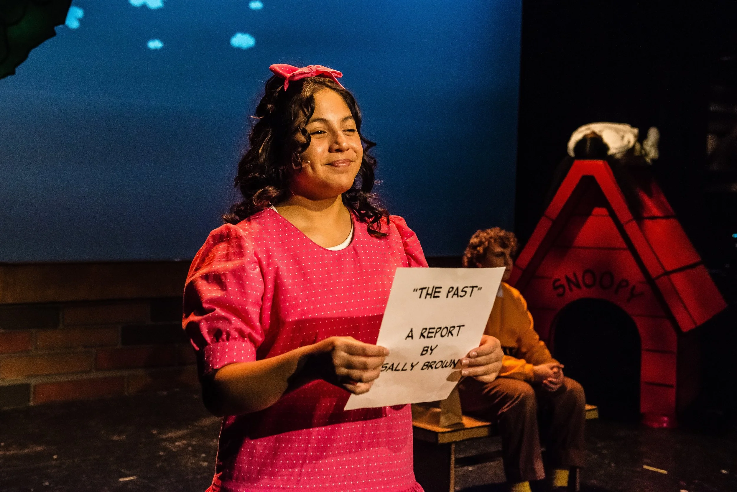 A woman playing Sally with curly brown hair, wearing a pink polka-dot dress and a pink bow, is holding a piece of paper that reads 'THE PAST'. There is a blue backdrop with clouds; Snoopy is lying on a doghouse in the background.