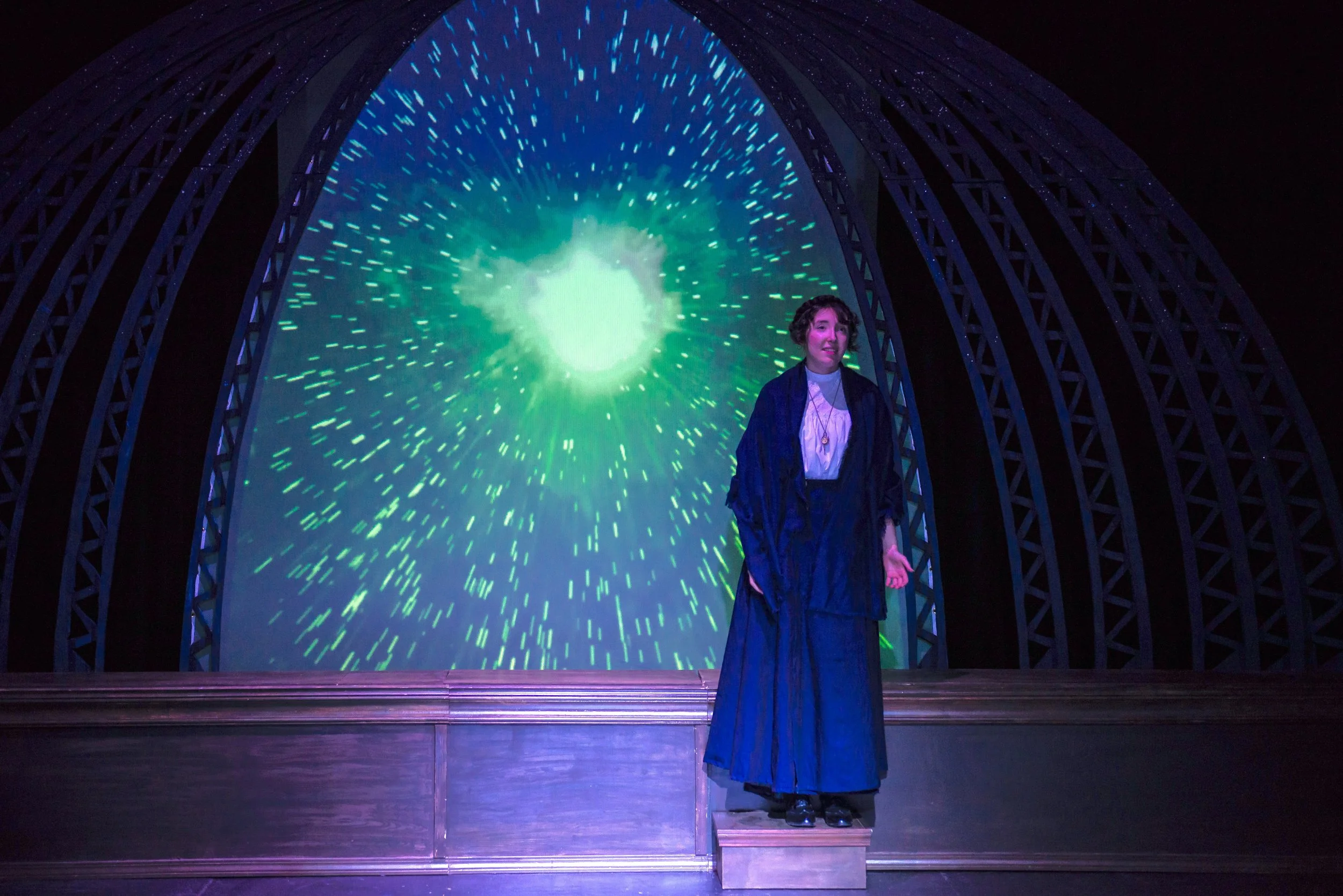 A woman standing on a stage for a production of the play 'Silent Sky' with a structural arch and a supernova display behind her.