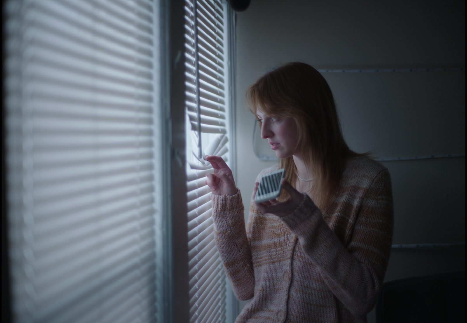 Young woman with red hair looking pensively out a window with blinds, holding a phone.
