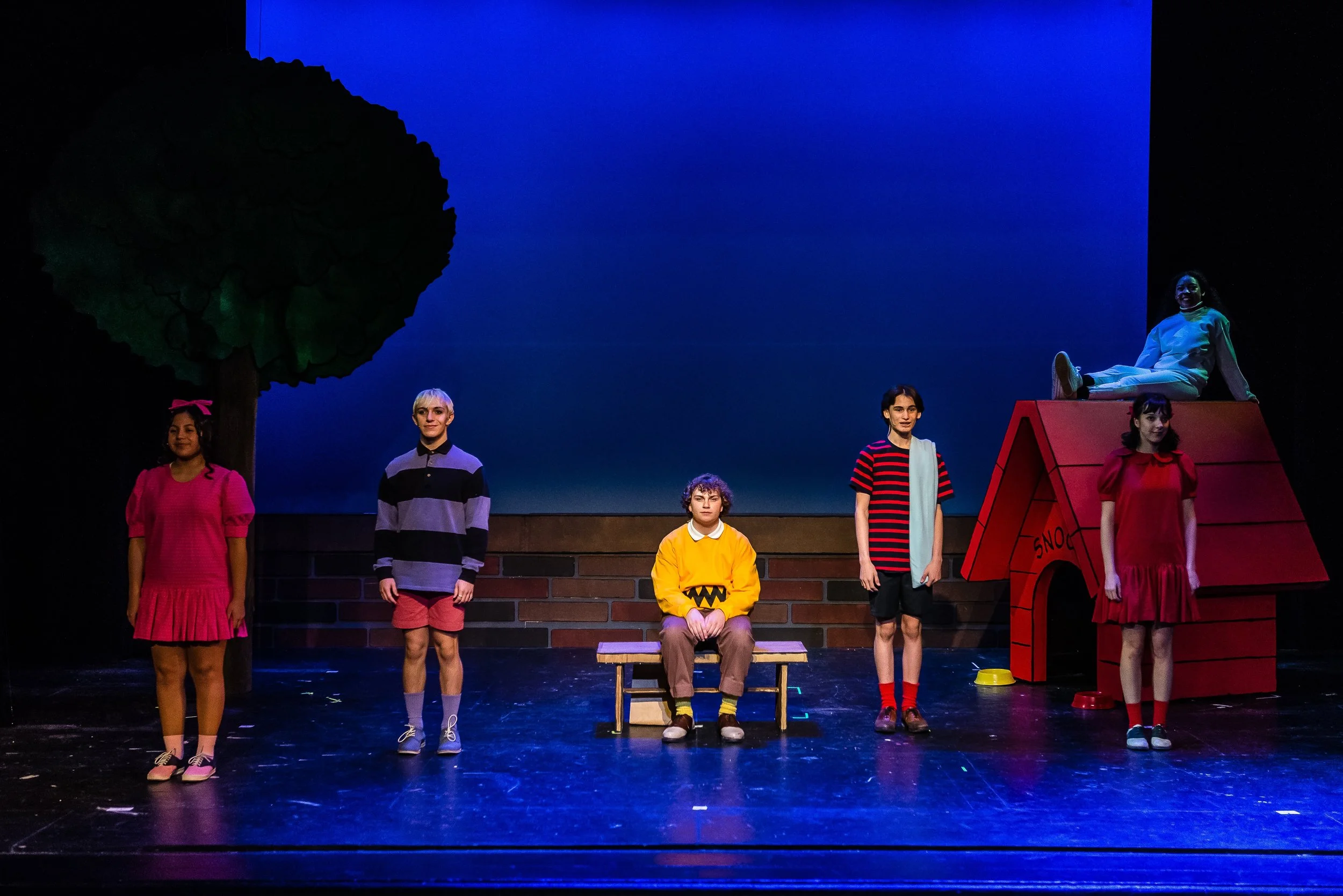 A group performing 'You're a Good Man Charlie Brown' in colorful costumes, with a dark backdrop and a large tree prop on the left, and Snoopy's red dog house on the right.