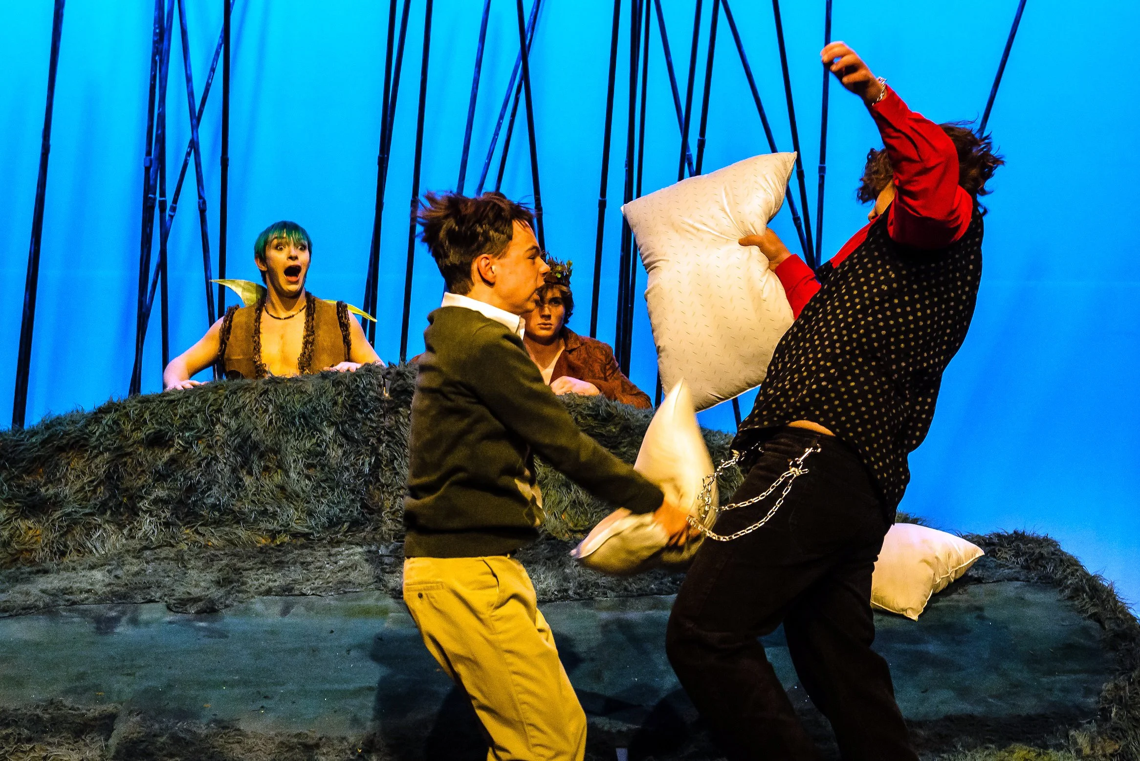 People in a pillow fighr during the performance of a play on stage with a blue background, including a man with curly hair holding a pillow, a man with short hair and a blazer, and Puck with short green hair, wings and a surprised expression.