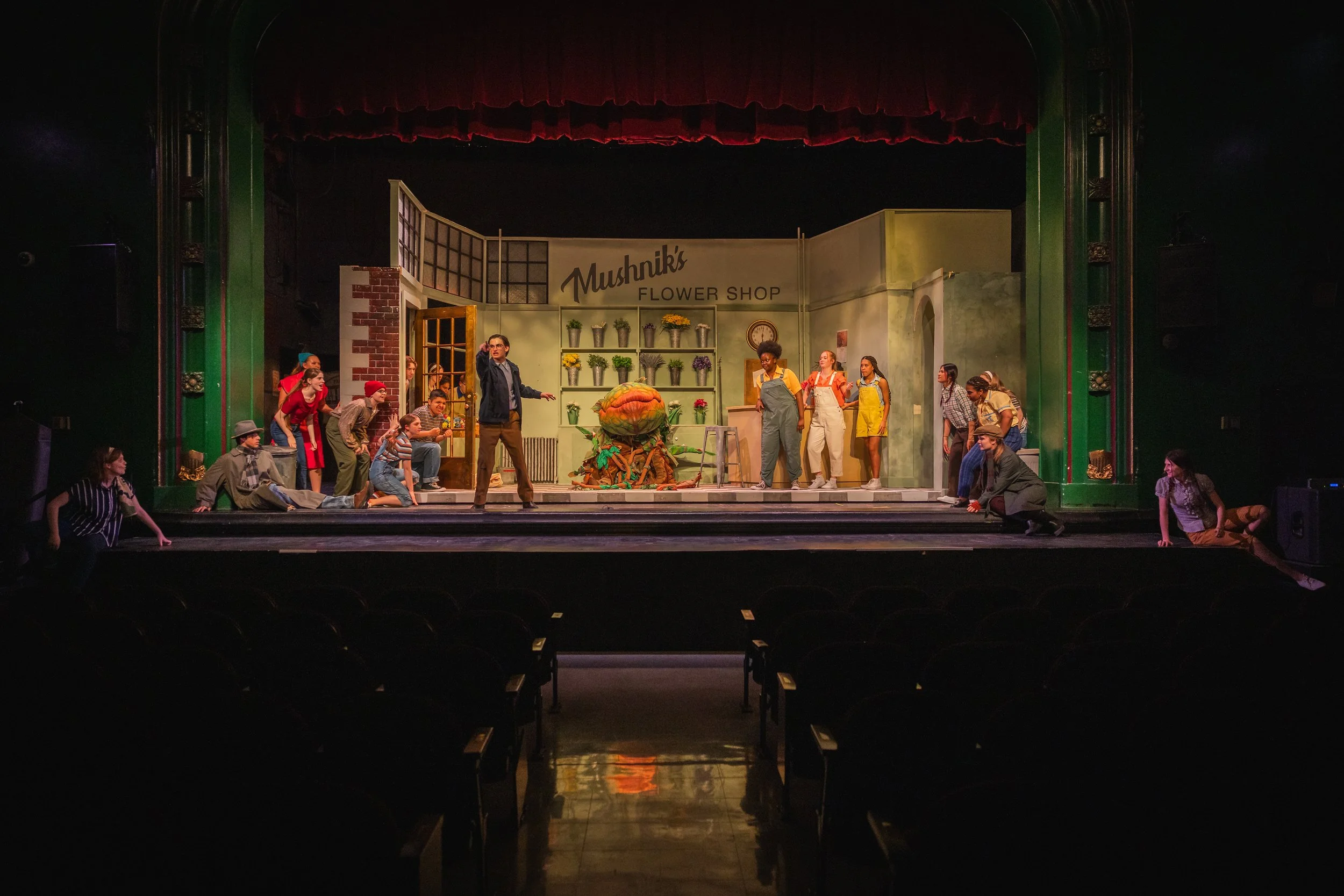 Stage scene from a theatrical production of 'Little Shop of Horros' set in 'Mushnik's Flower Shop' with actors in costume, set decorated with flowers and props, and a person speaking at center.