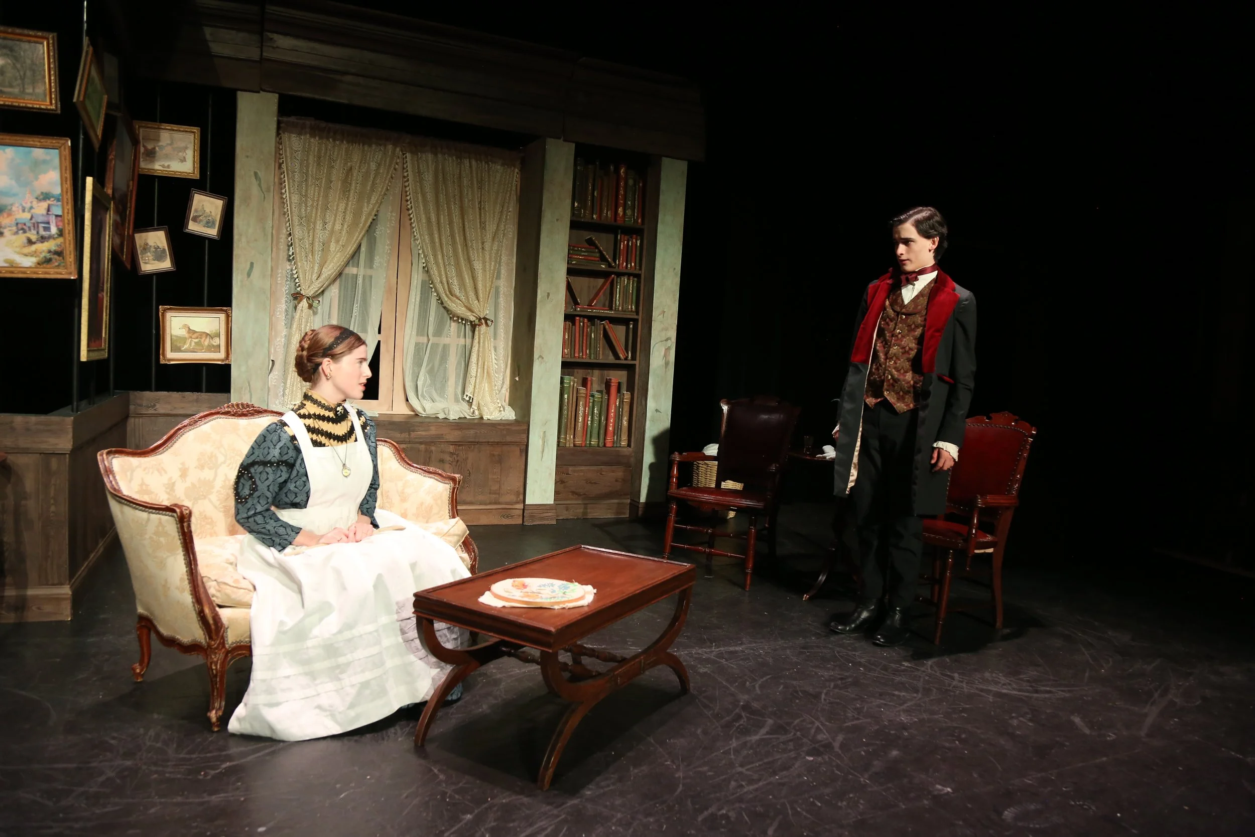 Actors performing a scene from a play set in a Victorian-style room with paintings, curtains, and bookshelves, depicting a woman sitting on a sofa and a man standing nearby.