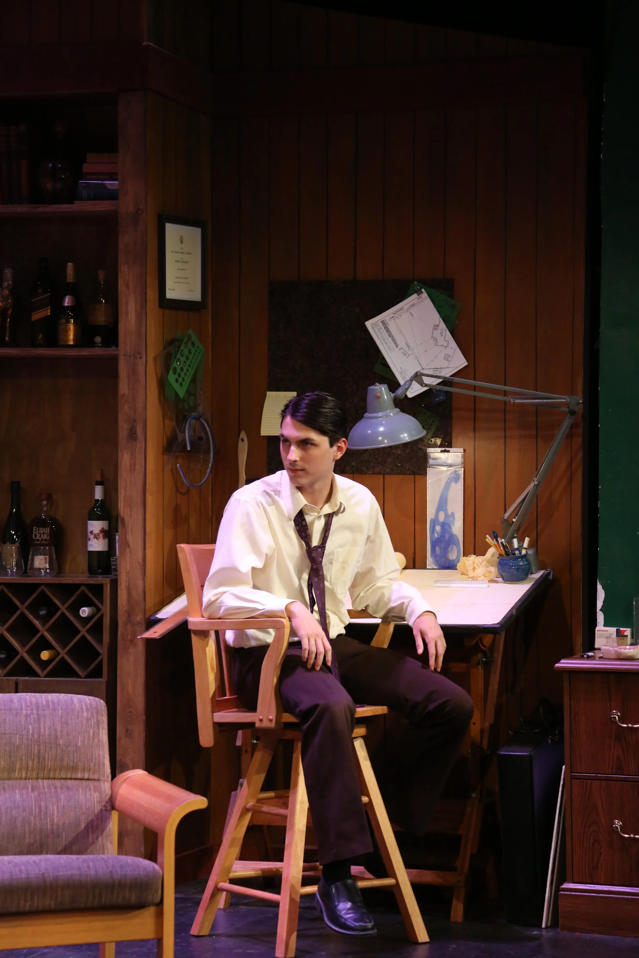 A young man with dark hair wearing a white shirt, purple tie, dark pants, and shiny black shoes is sitting at a wooden drafting table in a cozy room with wood-paneled walls and shelves with bottles and books.