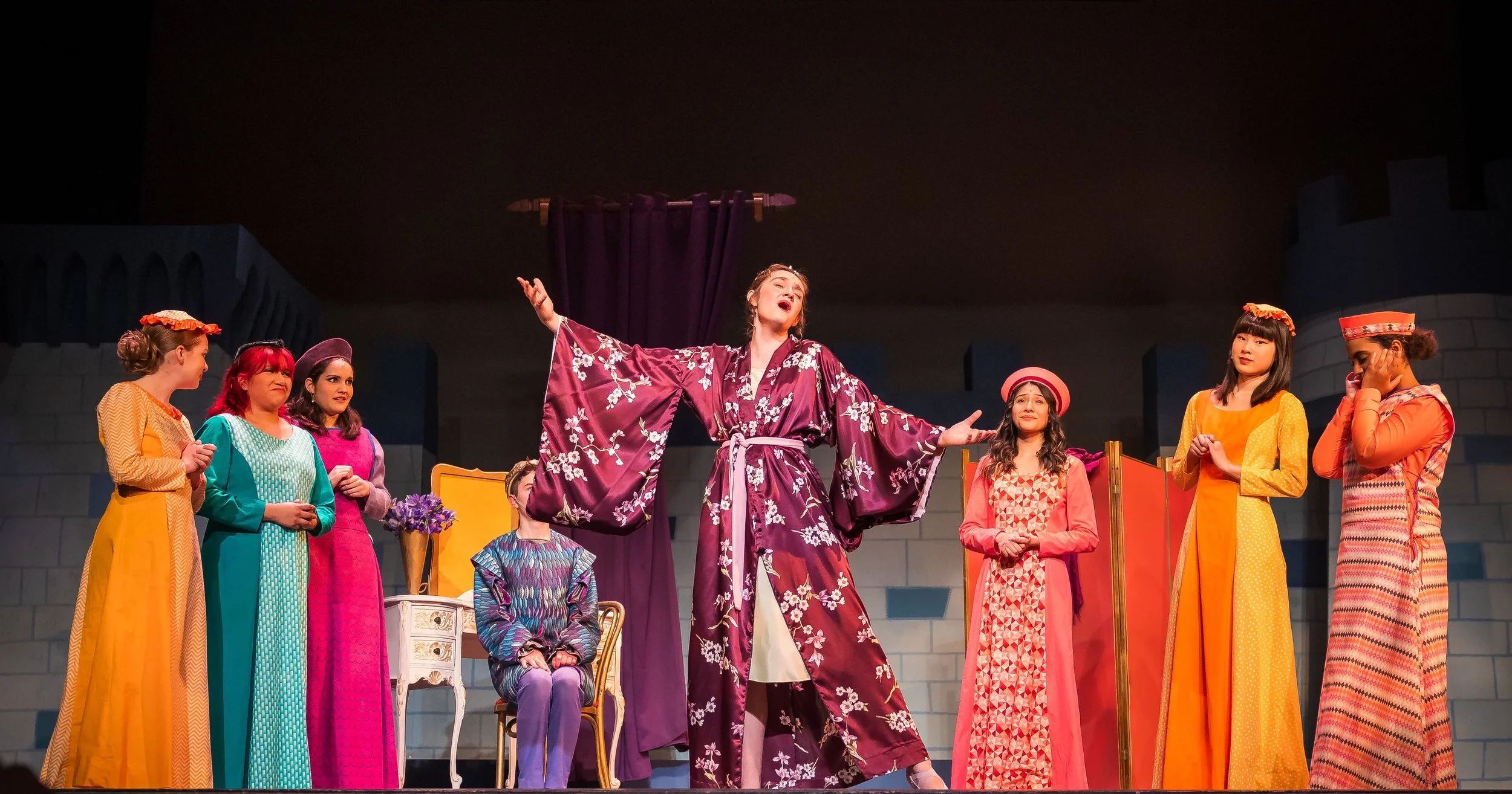 Theatrical performance with women dressed in colorful costumes on stage, with one woman in the center singing with arms outstretched, surrounded by others observing.