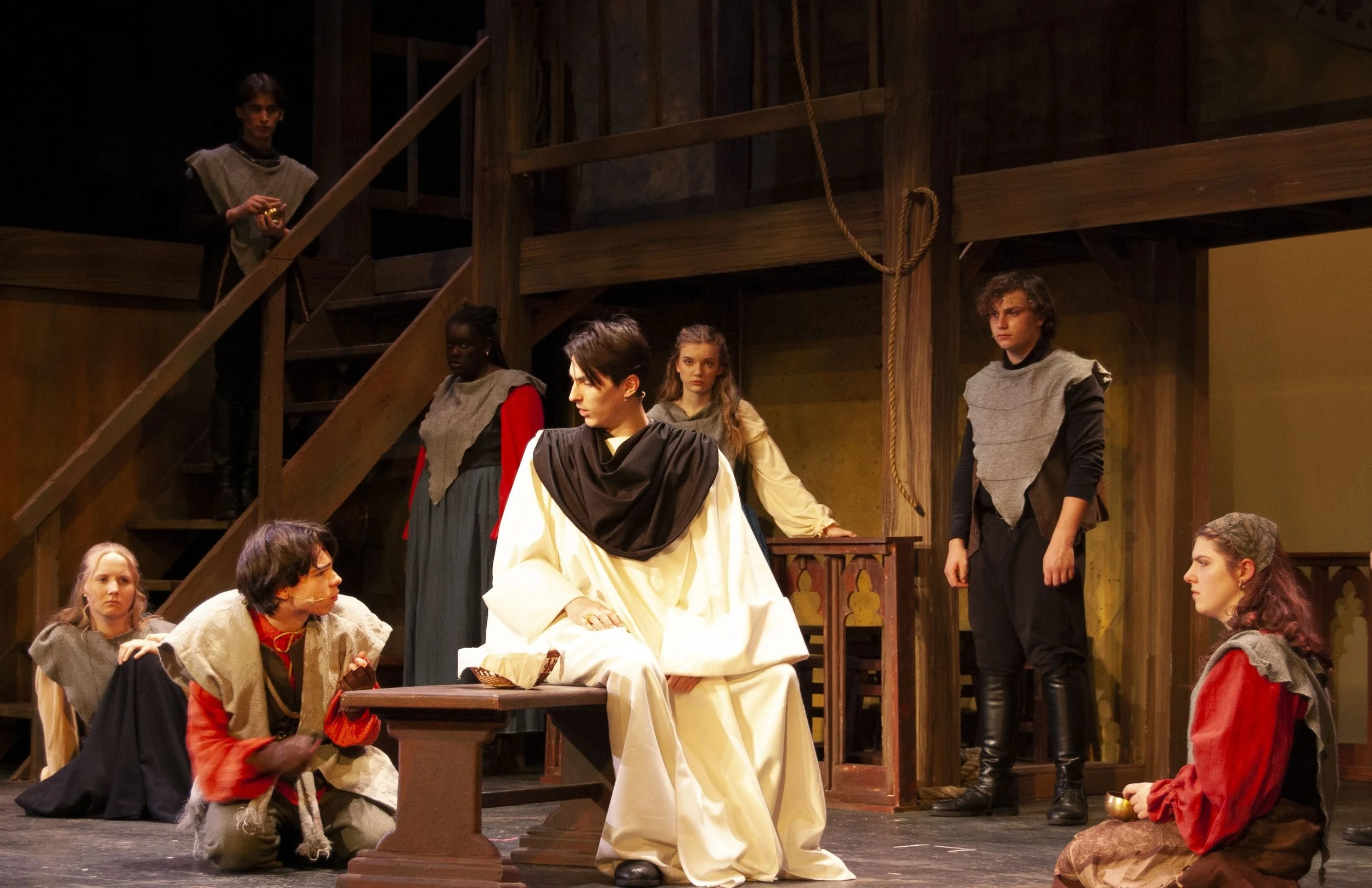 Actors on stage performing a play set in a medieval setting, with wooden stairs and ropes, characters dressed in period costumes, engaging in a dramatic scene.