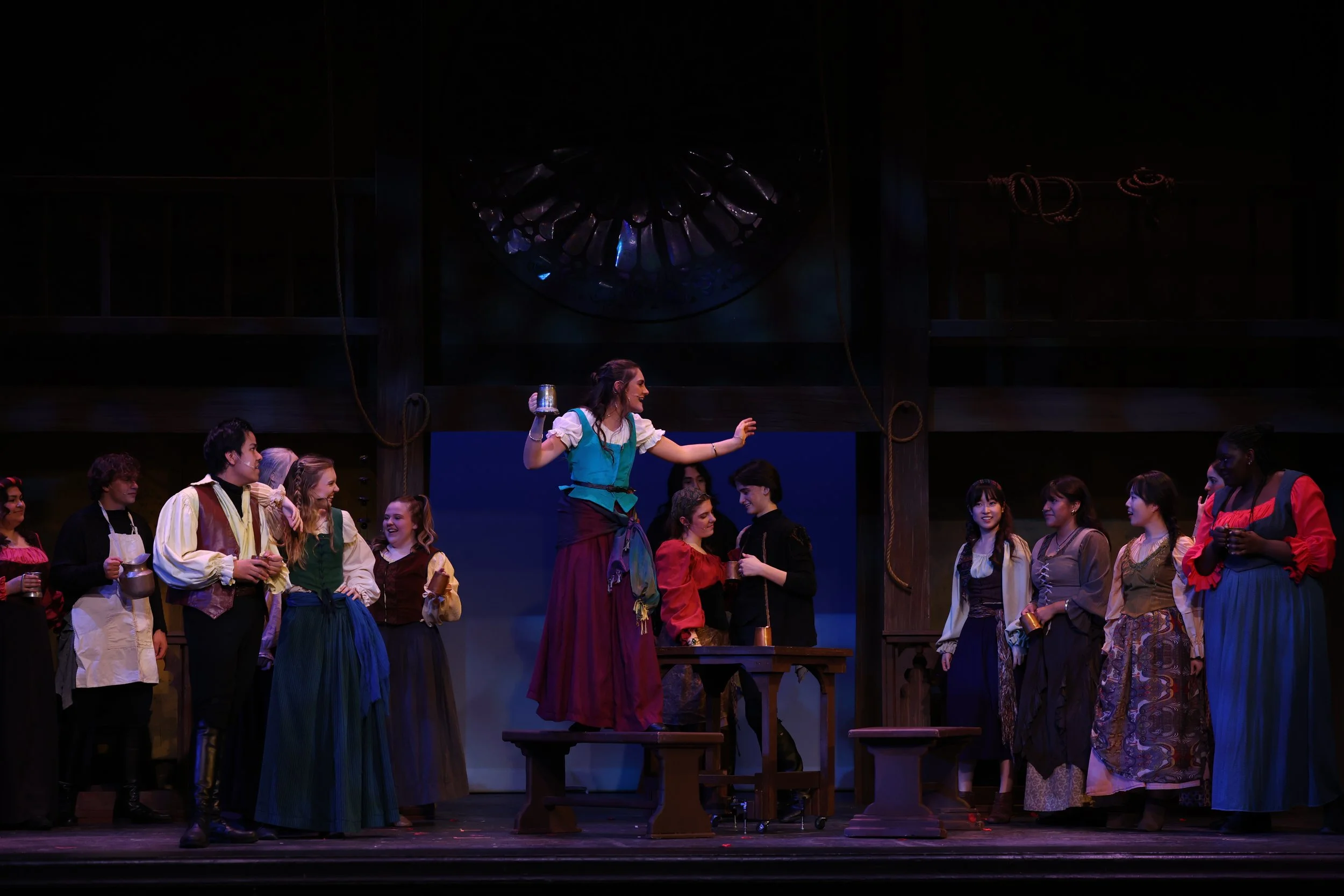 A group of people dressed in period costumes on stage, participating in a theatrical performance, with some holding goblets.