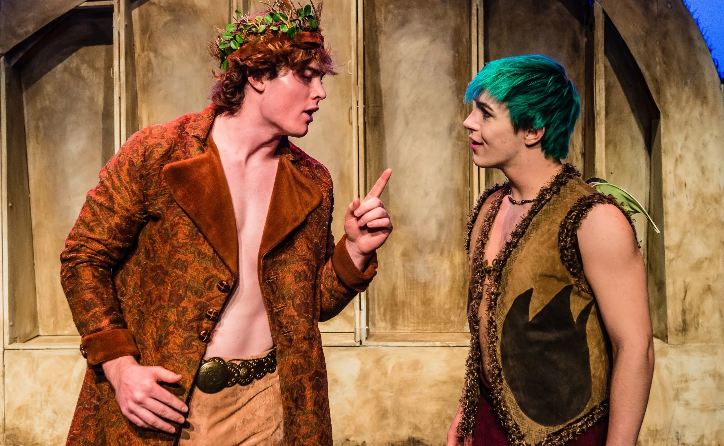 Two young actors engaged in a dramatic scene on stage, with one wearing a brown patterned jacket and the other in a brown vest with green and black hair, set against a wooden backdrop.