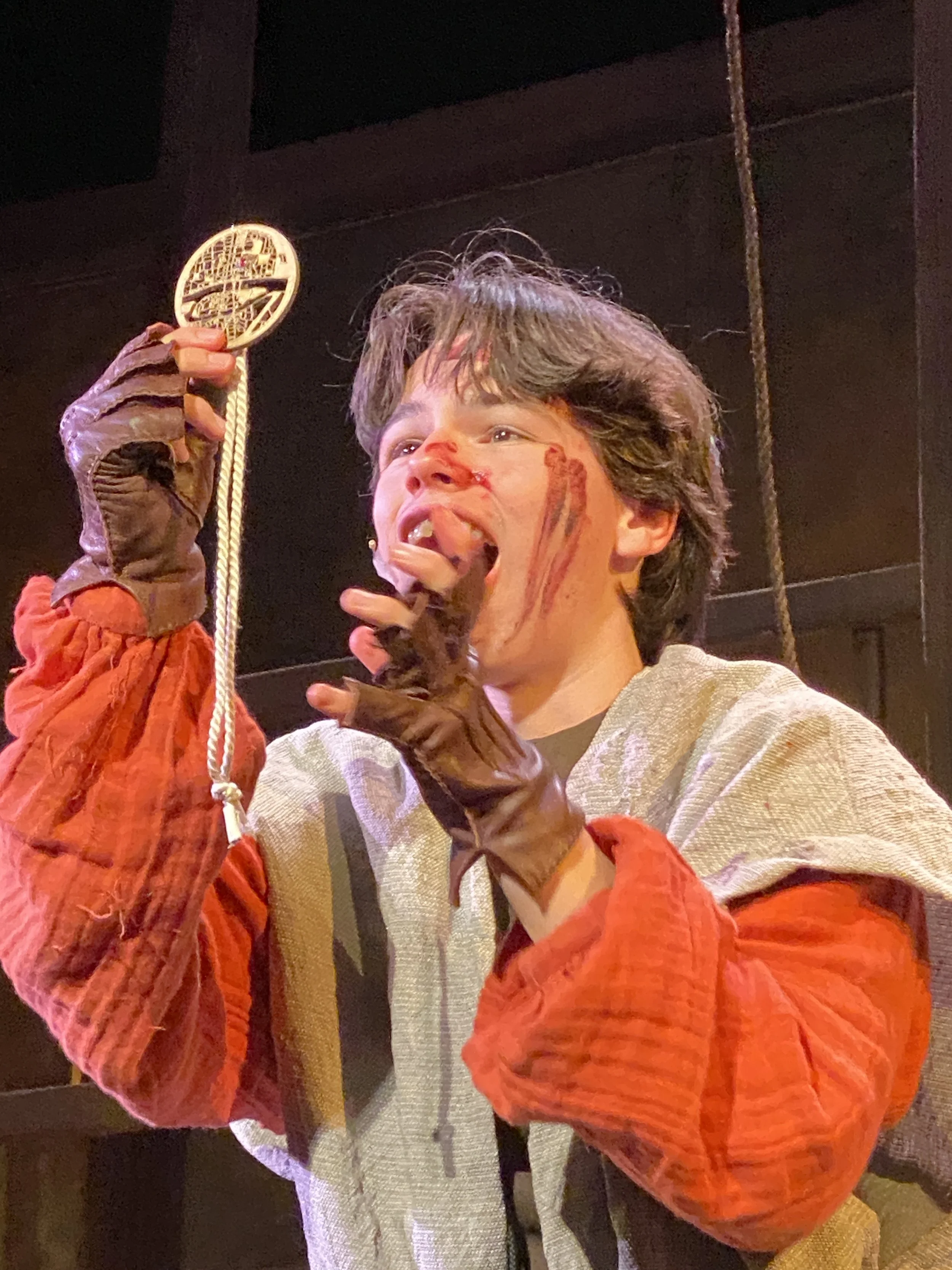 A person with short brown hair wearing a light-colored textured jacket, brown gloves, and an orange puffy sleeve, is holding a round, flat object with intricate cut-out designs on a string. Their face has red makeup resembling blood.
