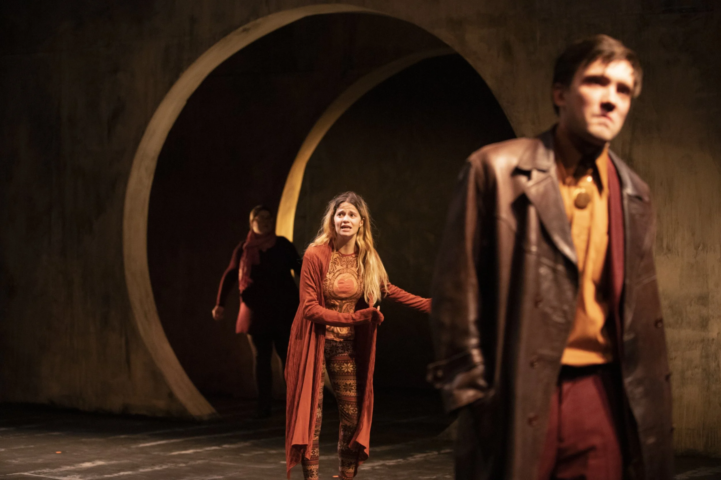 The image depicts a stage scene with four people. A woman with long, wavy hair, wearing a patterned outfit and an orange cardigan, has an expressive look on her face as she gestures with her hand. Two men, one with short dark hair, wearing a leather 
