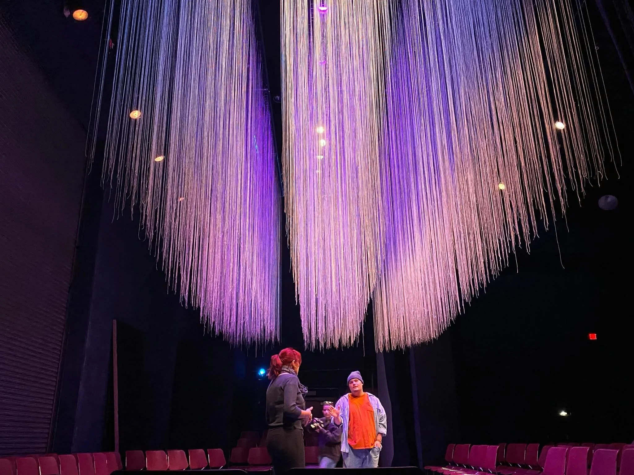 A stage production with people standing and talking underneath a large, colorful installation of hanging strings in purple, pink, and white, inside a dark room with purple chairs.