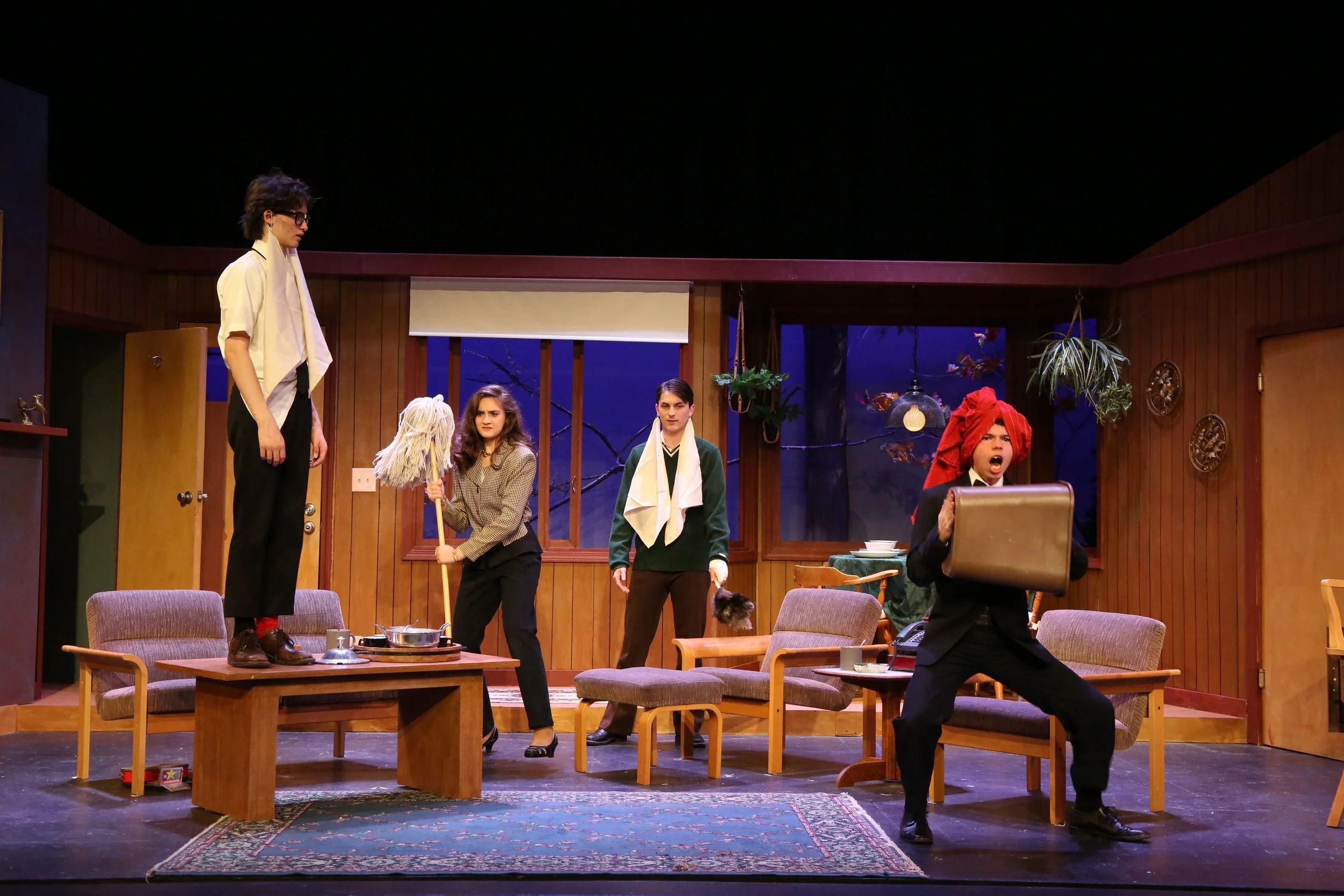 Stage scene from a play involving four actors and various props, with a wooden backdrop, plants, and a blue sky visible through the windows.
