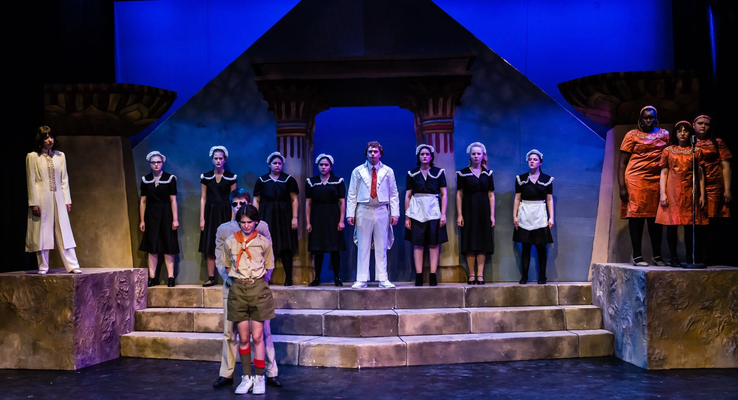 The image shows a theatrical stage with multiple performers in costume. Some are dressed as nuns in black and white outfits, and others are in orange dresses with headbands. There are two characters in front, one in a tan outfit with glasses and anot