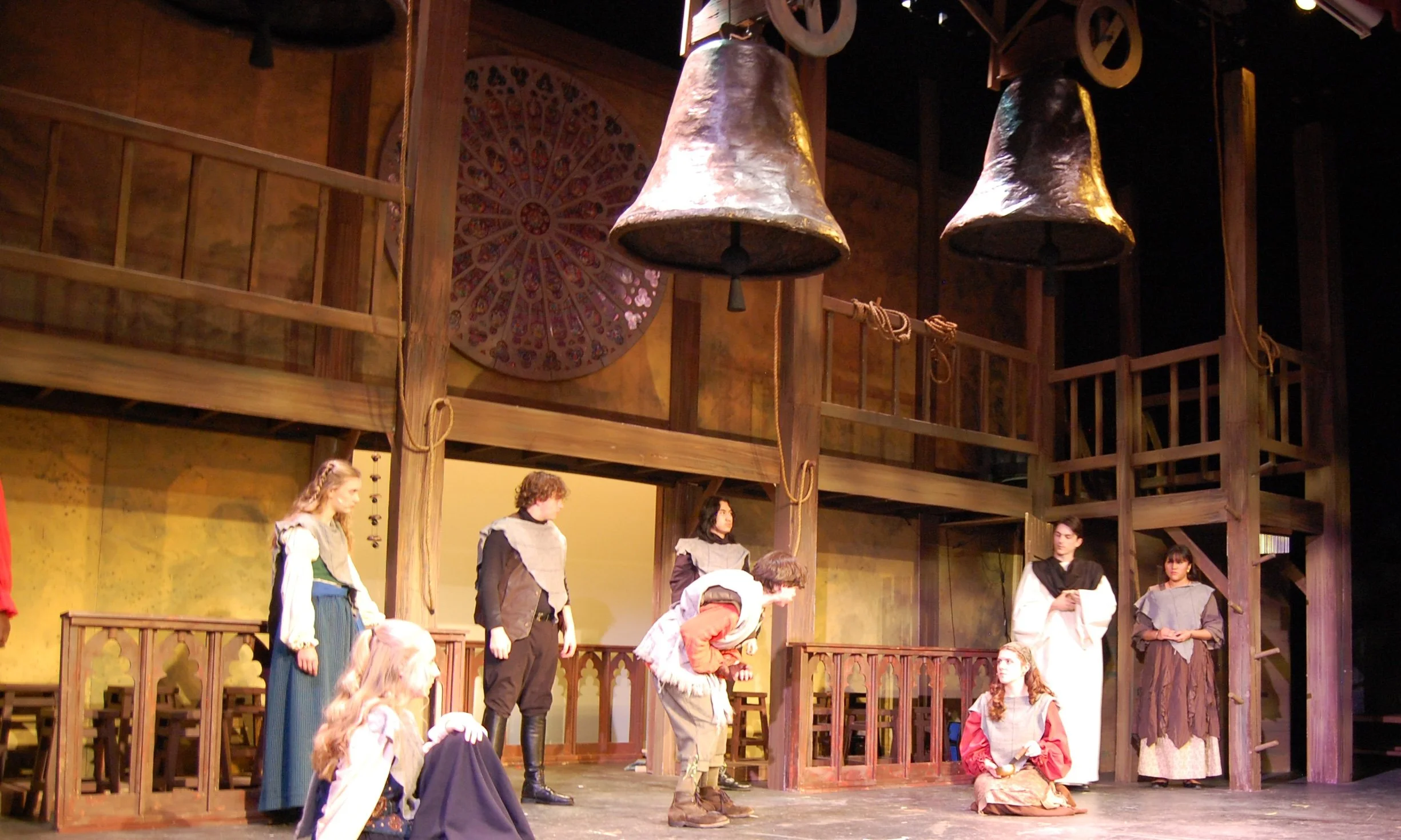 People dressed in medieval costumes preparing for a stage performance on a wooden set resembling a cathedral, with large hanging bells and a colorful circular rose window in the background.