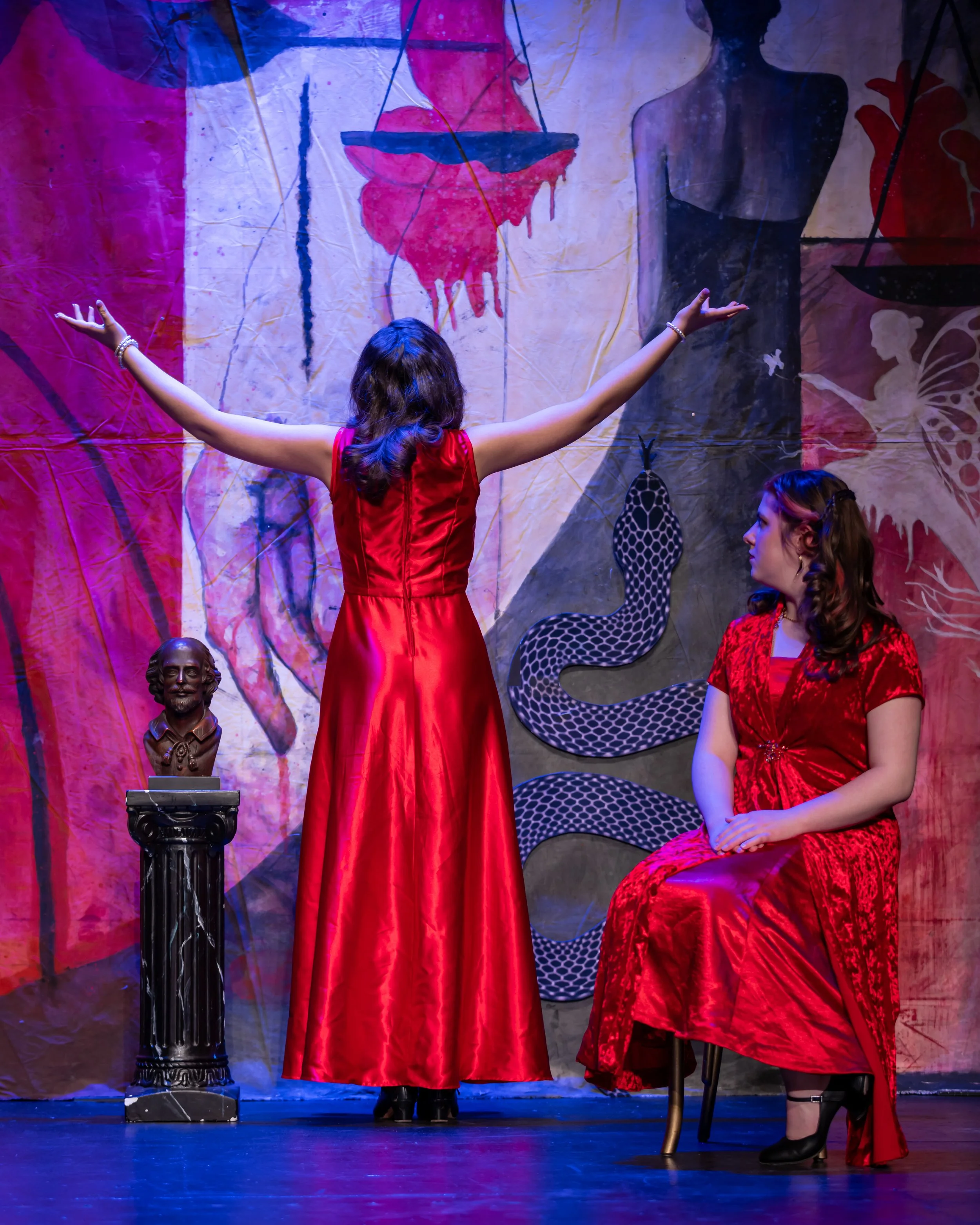 The image shows a scene from a stage play featuring two women in red dresses. One woman, with her back to the camera, stands with her arms outstretched, facing a colorful backdrop with a silhouette of a woman and various patterns. The other woman is 