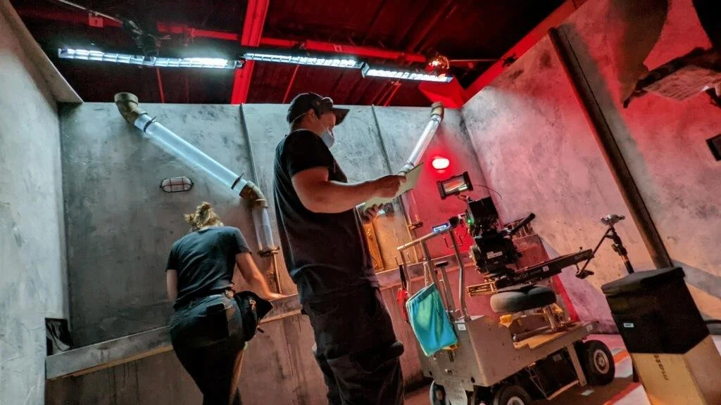 Two workers in a sound stage, one reading a paper and another working at a table, with exposed pipes and red lighting on the walls.