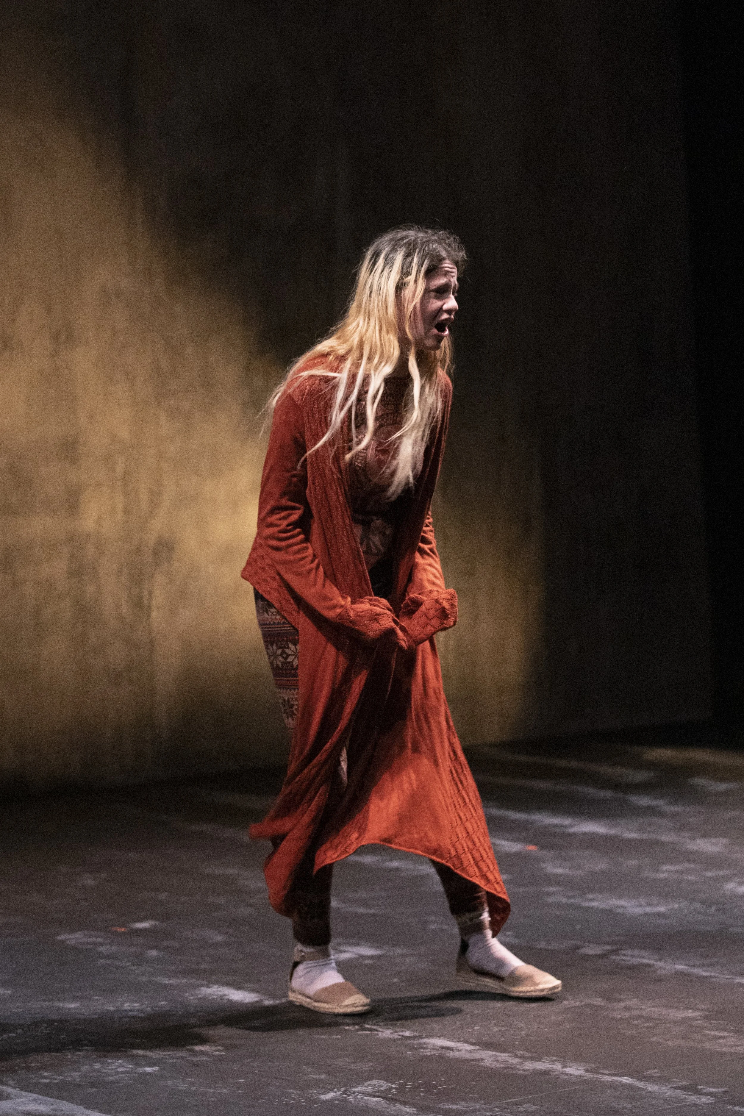 A woman with long blonde hair in a reddish-brown costume performing on stage. She is in motion with a dramatic expression. The background is a weathered, ochre-colored concrete wall.