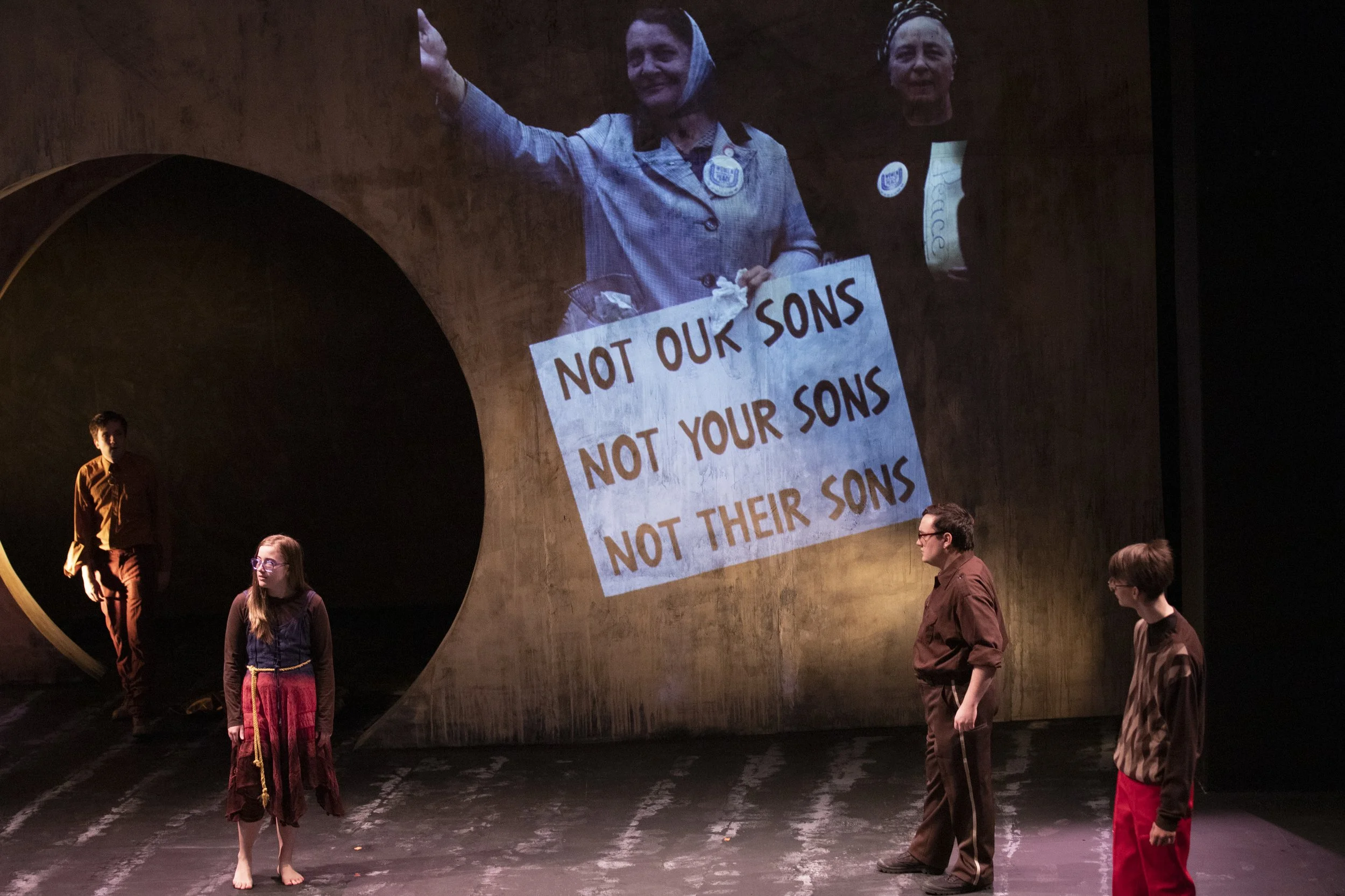 A group of people standing on a dark stage with a large projected image of a smiling woman holding a protest sign that reads 'NOT OUR SONS NOT YOUR SONS'. The scene is indoors, and some people are looking at each other while others are looking away.