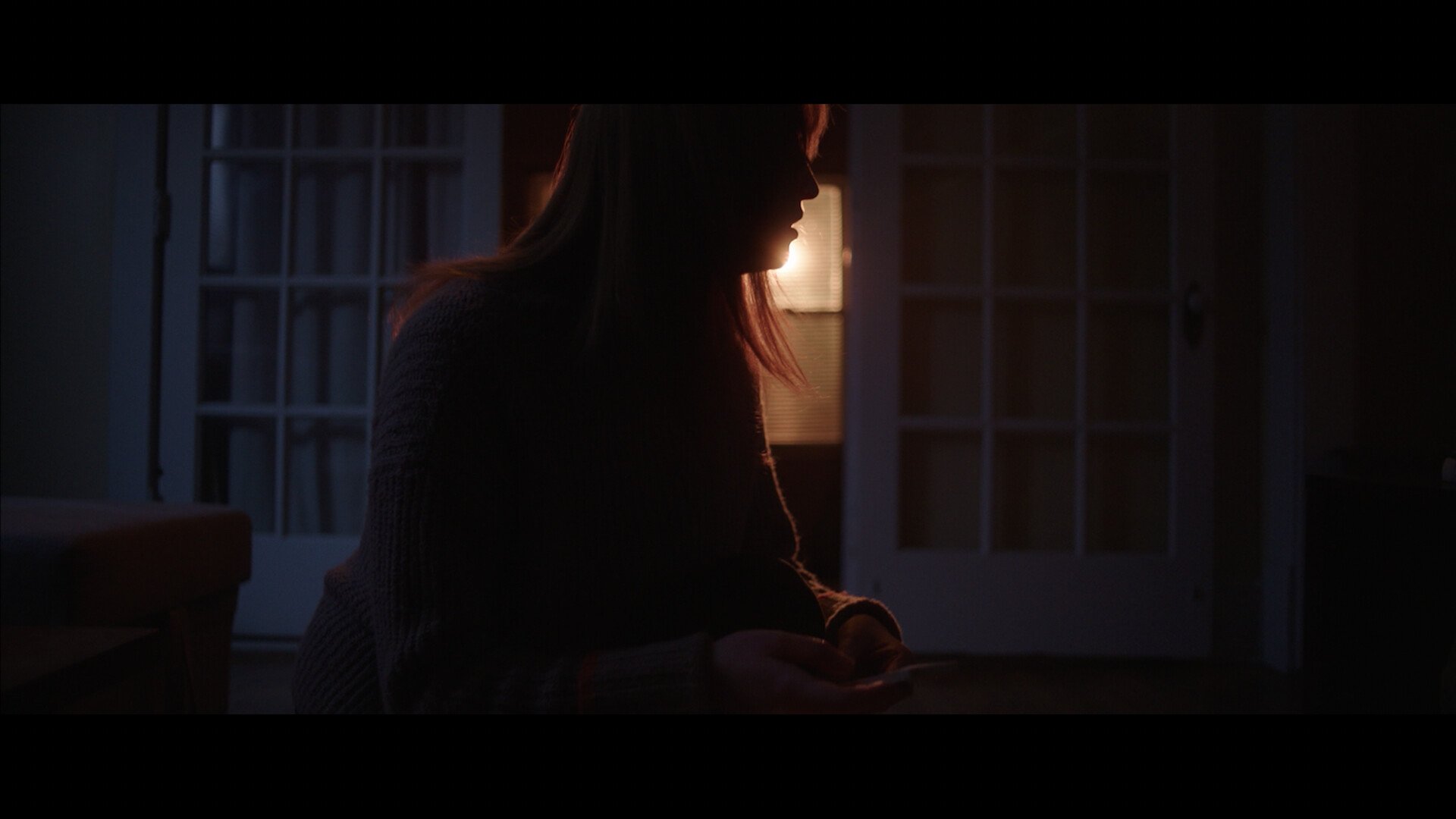 Profile of a woman with reddish hair sitting indoors in a dimly lit room, looking down, with a warm backlight from a window or door behind her.