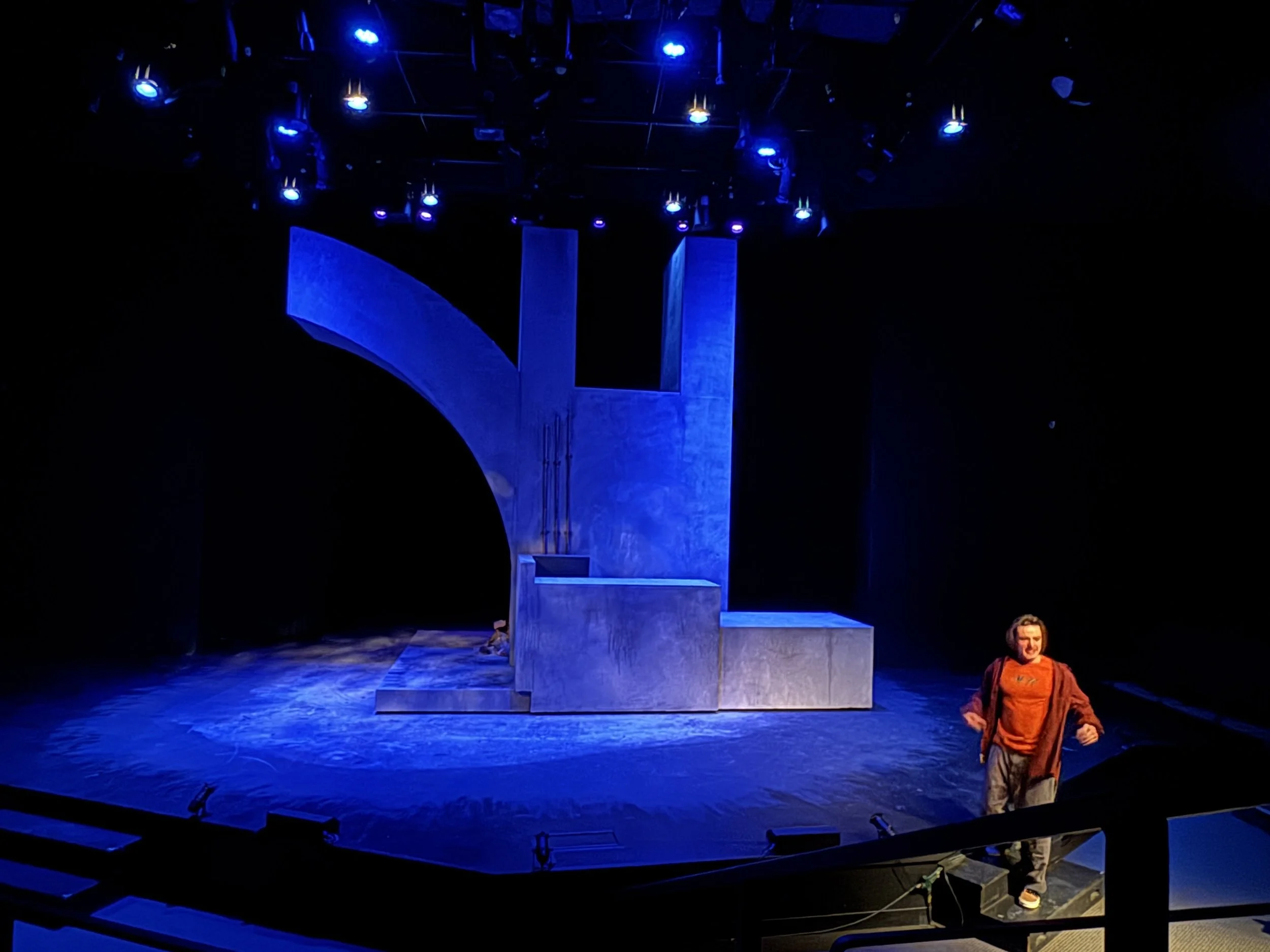 A dark theater stage with blue lighting, featuring minimal set pieces and a person in a colorful costume standing on the right side.