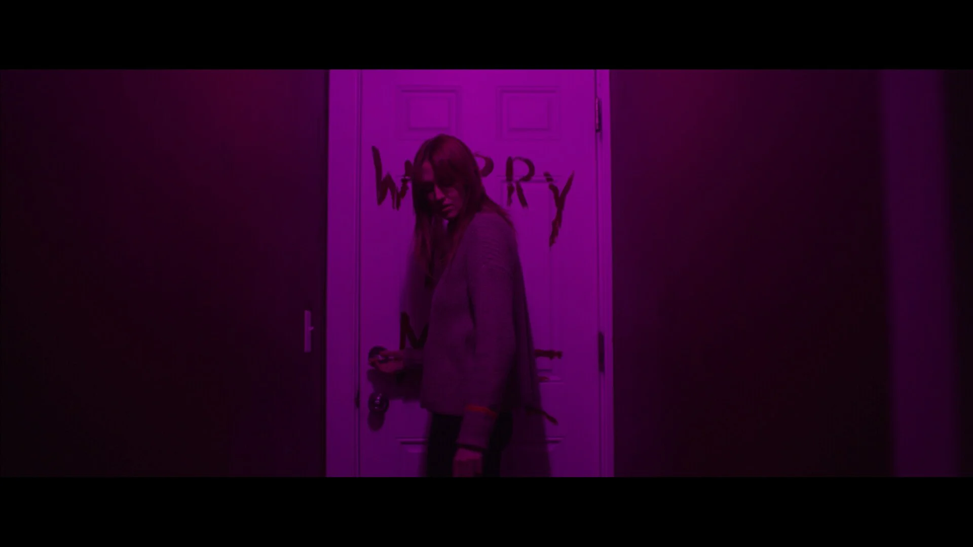 A woman standing in a dimly lit hallway illuminated with purple neon light, holding a phone, with graffiti on the door behind her.