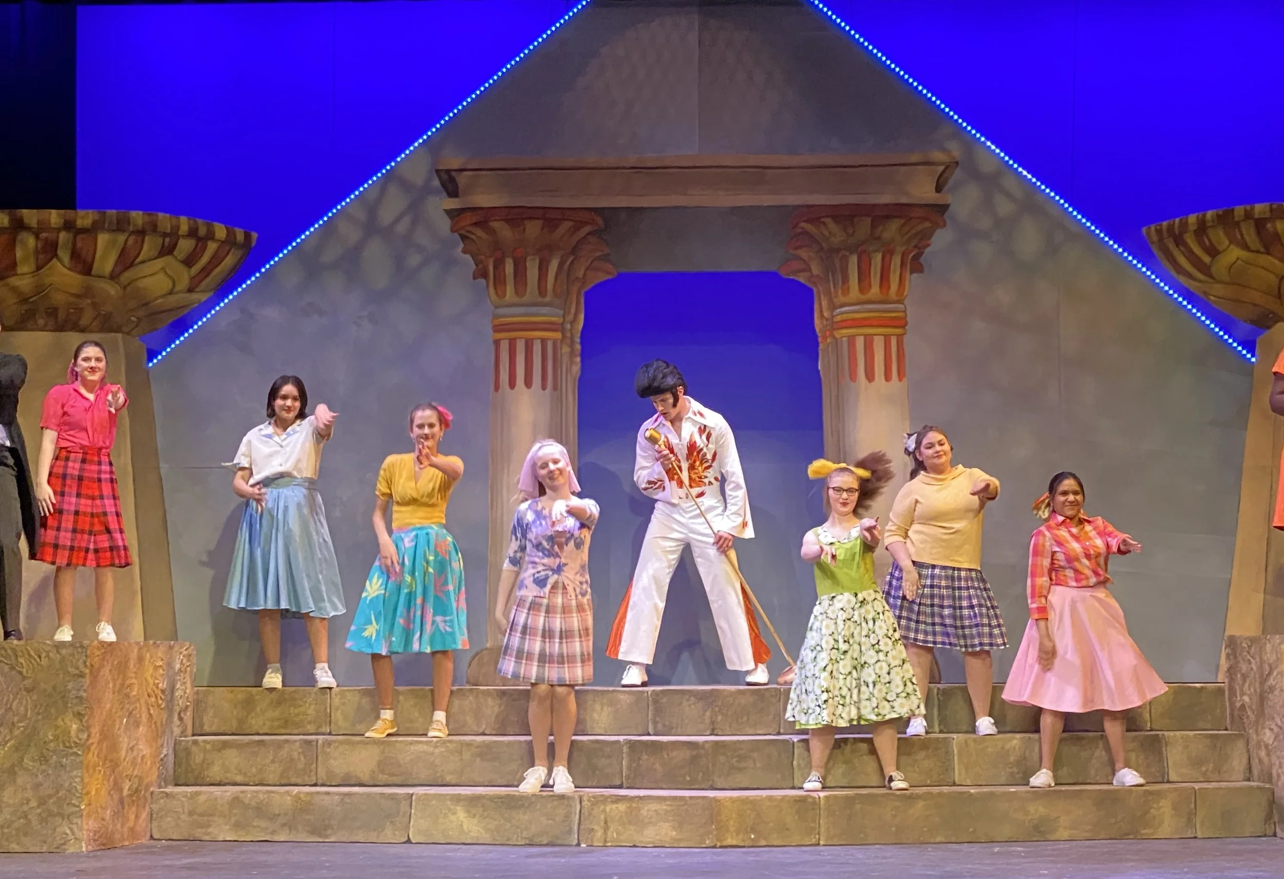 A group of performers on stage dressed in colorful retro clothing, with a set designed to look like ancient Egyptian architecture, including columns and a pyramid decorated with blue LED lights.