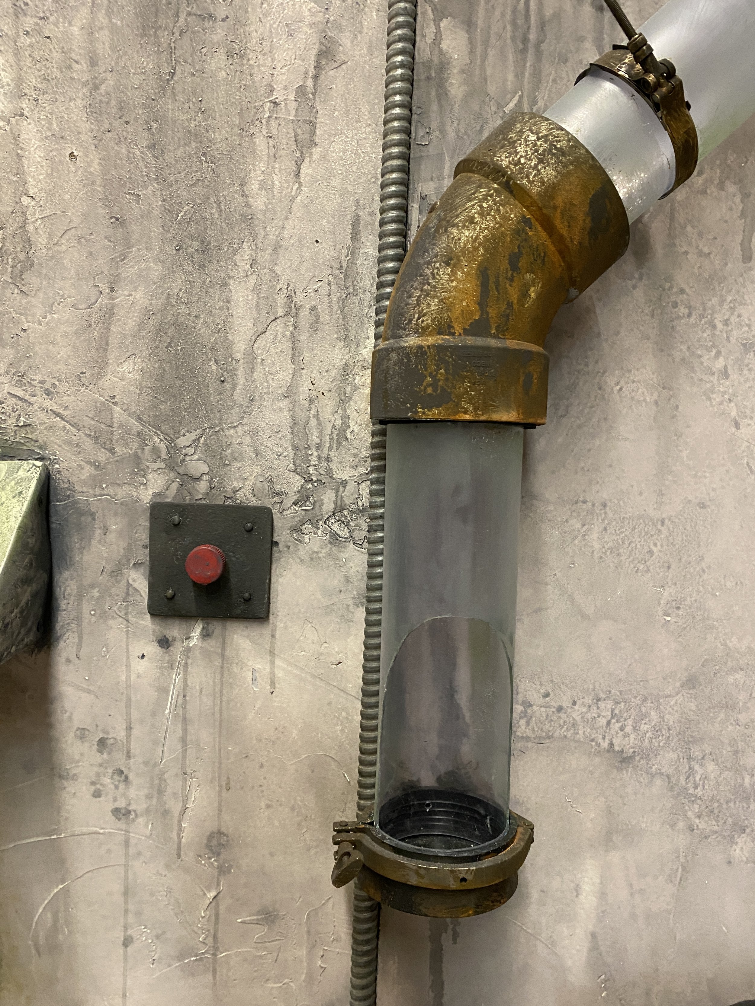 A metal pipe fitting with a flange at one end, mounted on a wall with visible wiring and a red button.