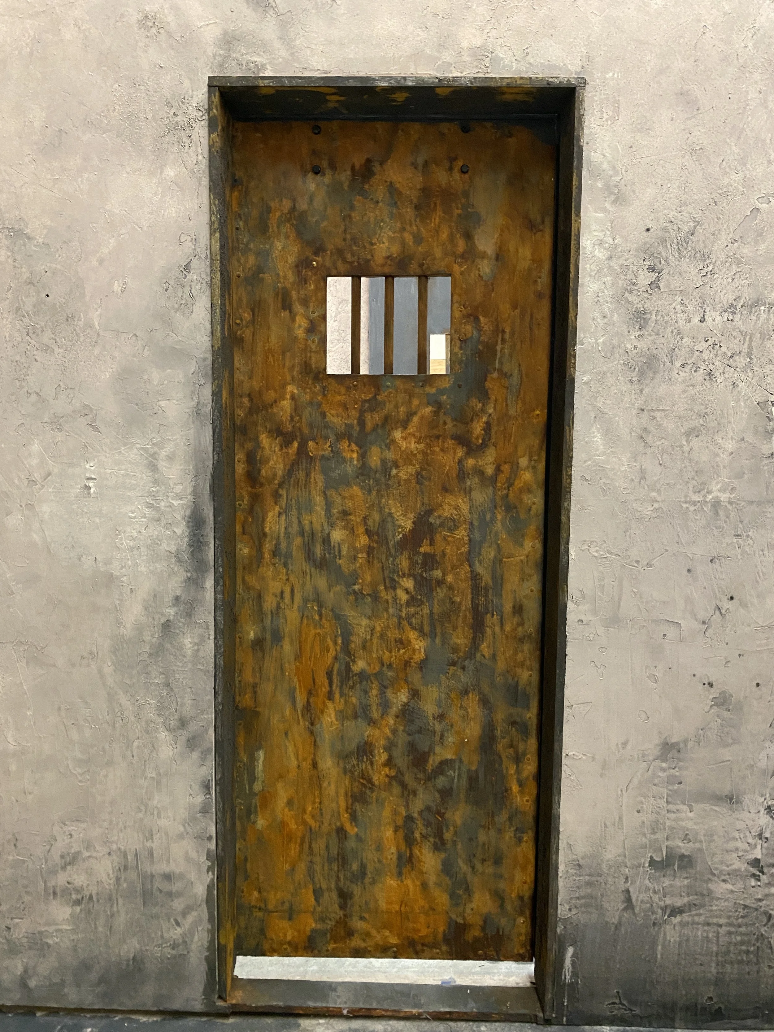 A film set of a rusty metal prison cell door with a small, barred window at the top, set within a concrete wall.