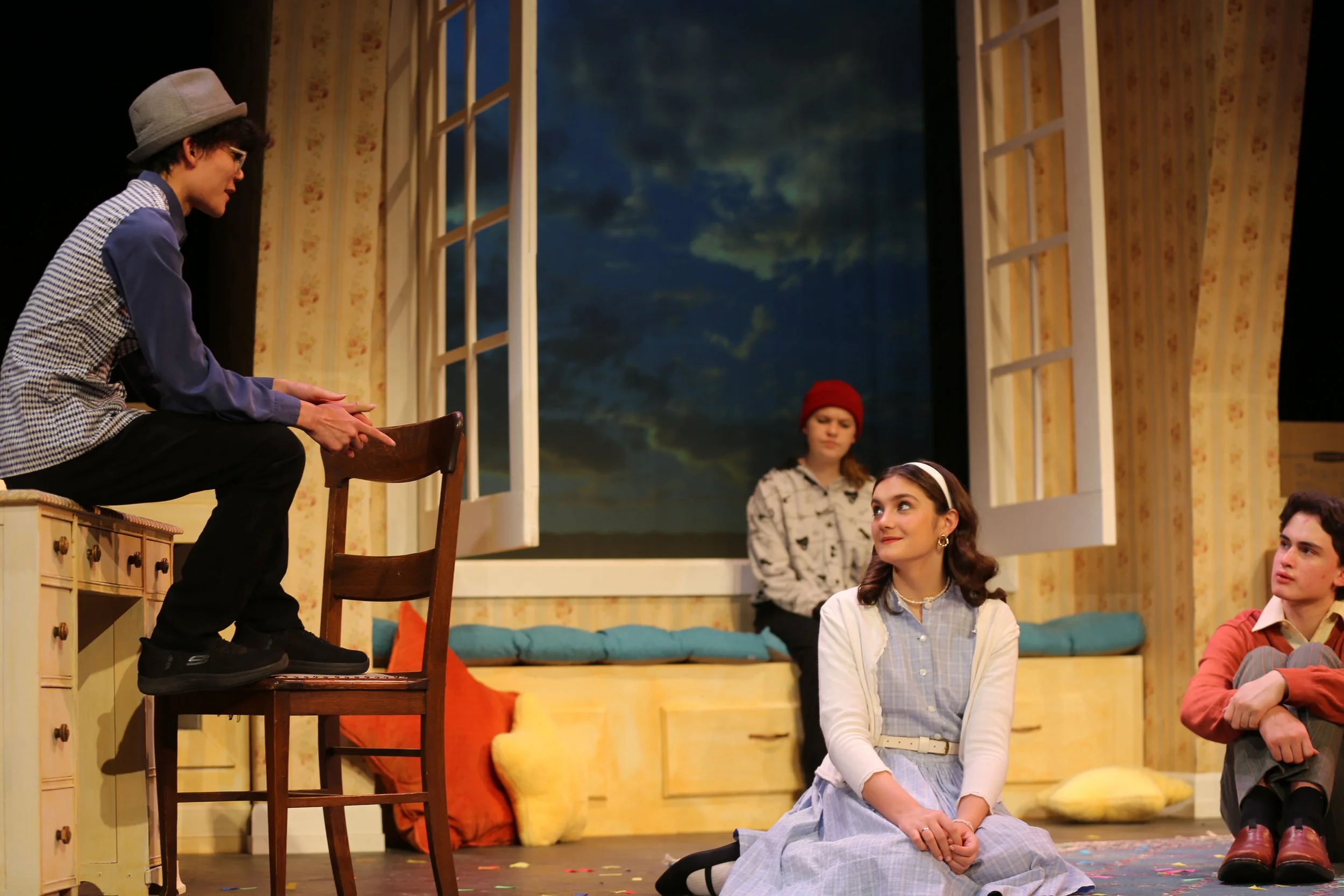 Group of young actors on stage during a theater performance, with one person sitting on a chair at a higher level, three people sitting on the floor, and a person standing in the background, in a room with floral wallpaper and a large window backdrop