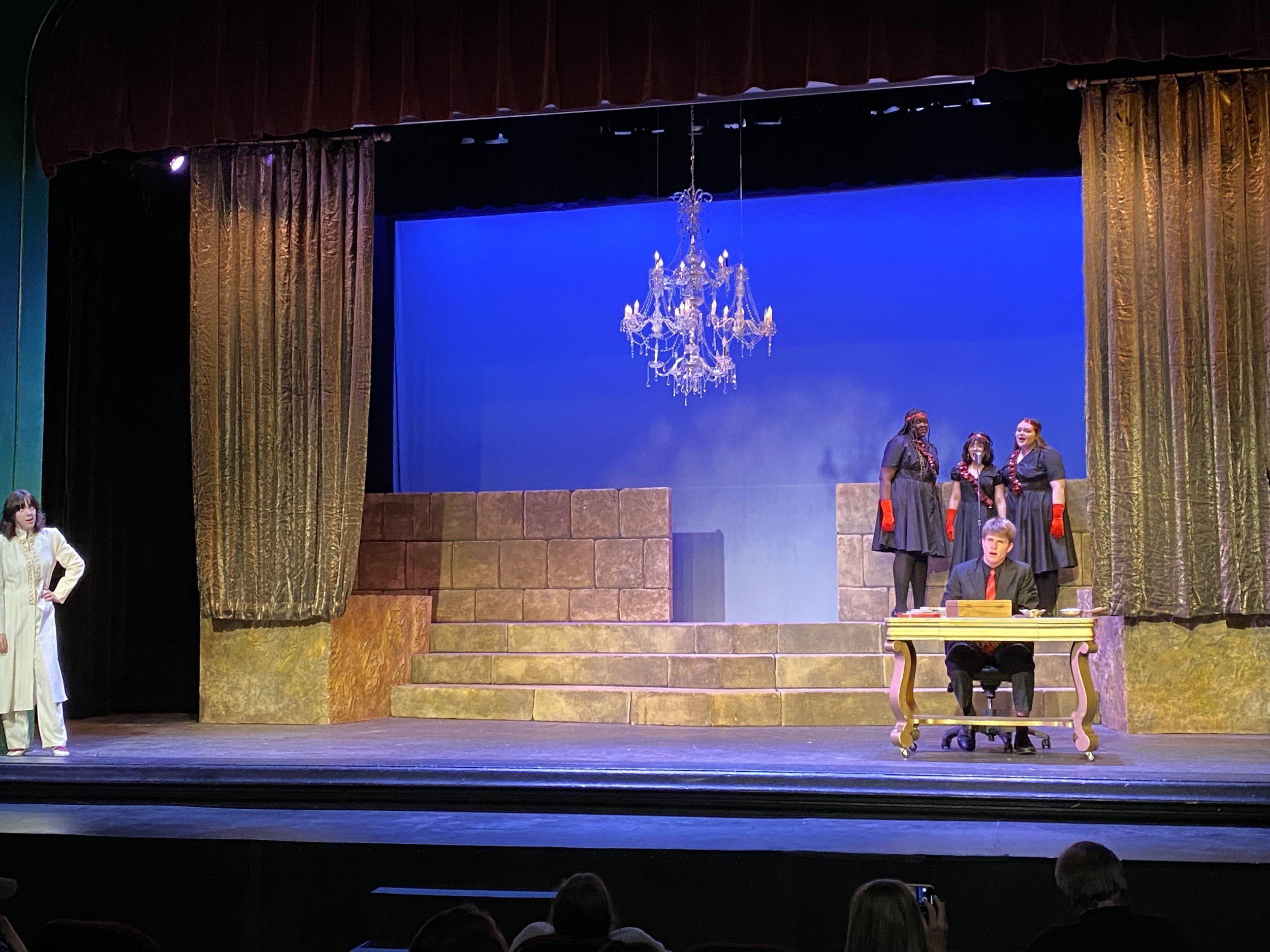 The stage of a theater show with gold curtains on both sides and a chandelier hanging from the ceiling. On stage, there are four women in black dresses with red gloves standing in a group, and a man sitting at a table with papers in front of him. A w