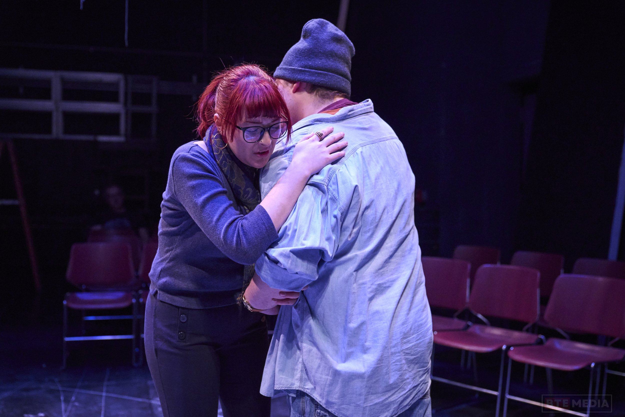 A woman with red hair, glasses, and a scarf hugging a man wearing a gray beanie and a light-colored jacket on a stage with empty chairs in the background.