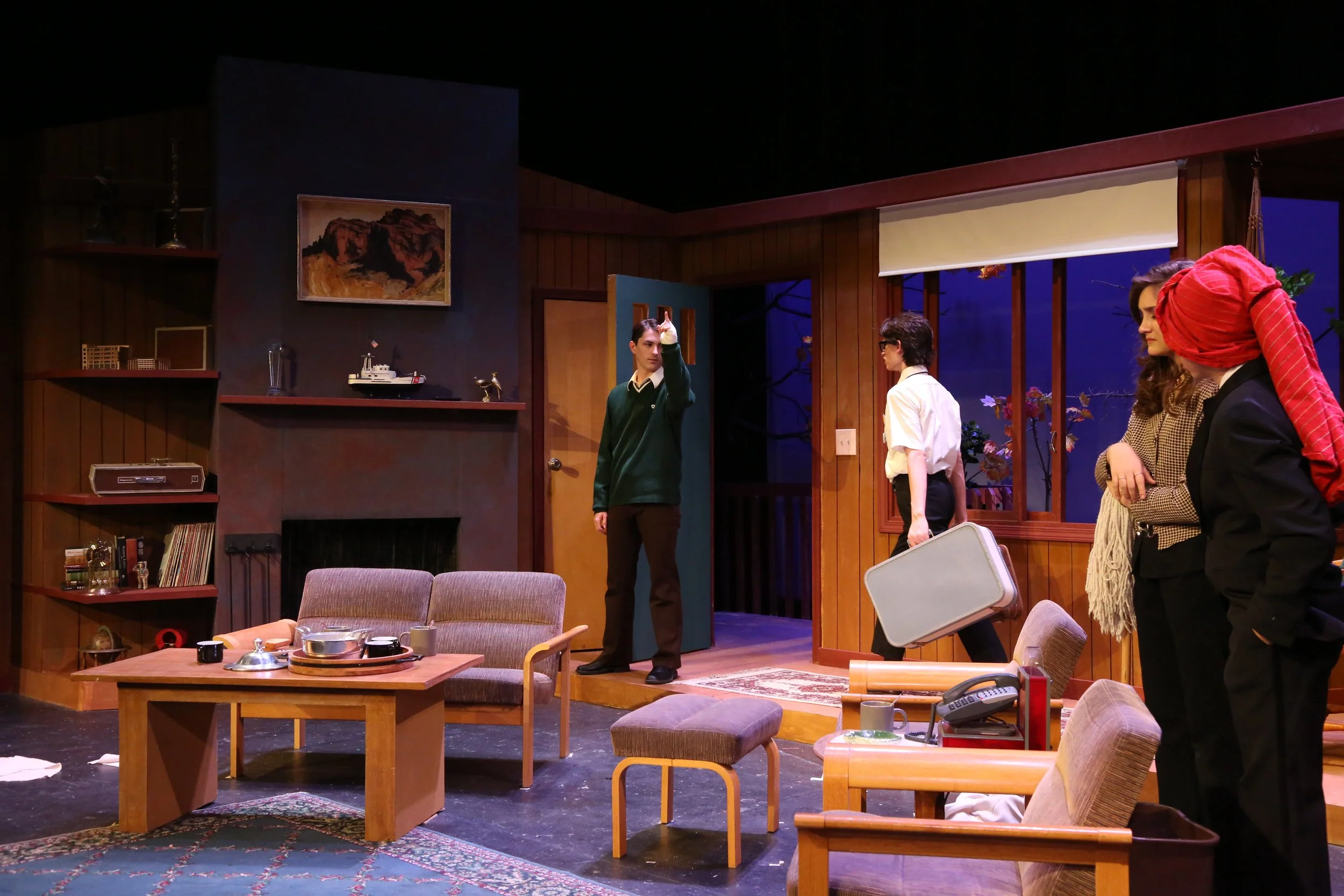 A scene from a theatrical play set in a cozy living room, featuring four actors on stage with wooden walls and shelves, a painting, and a window with a view of a blue evening sky.