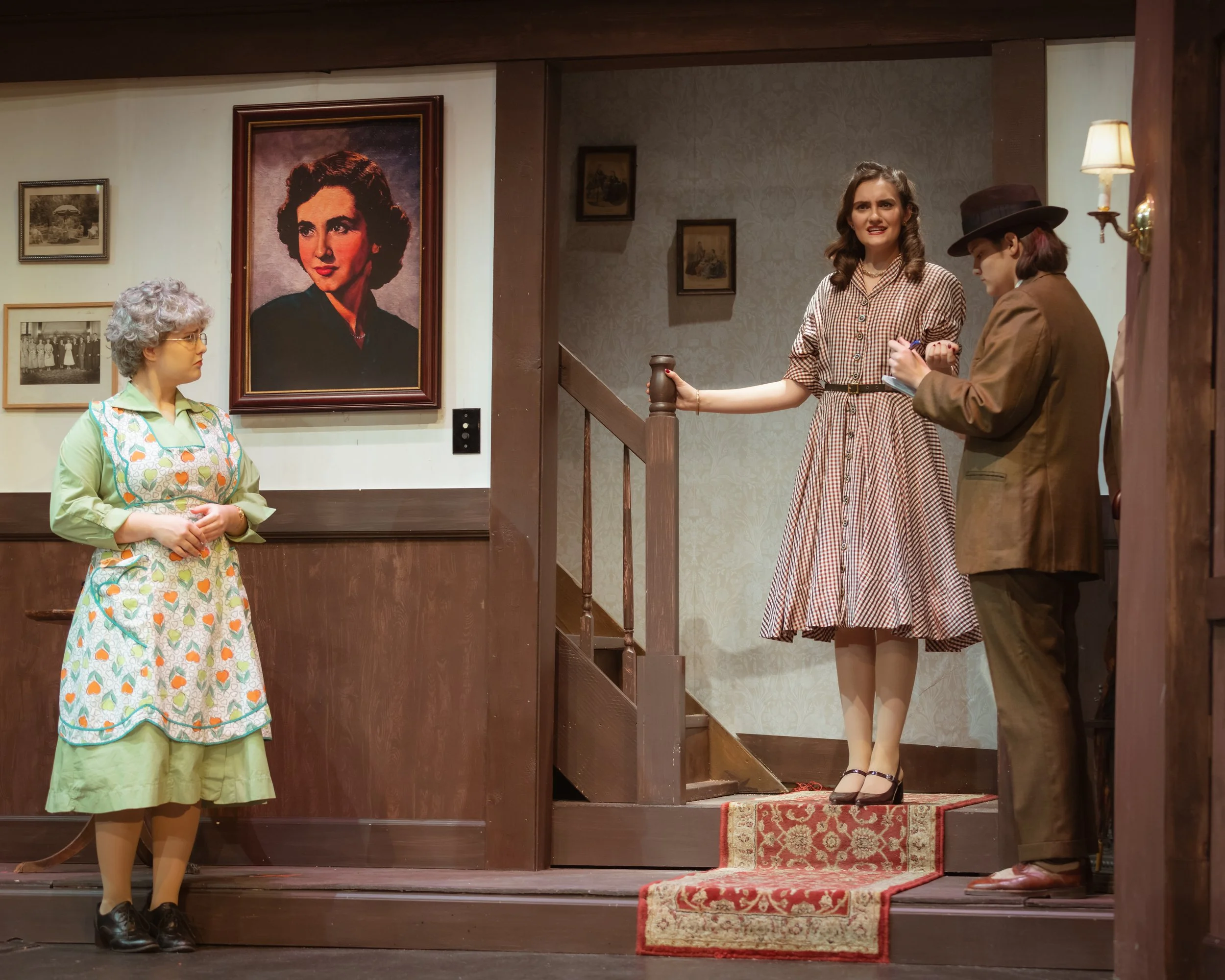 Scene from a theatrical play with three women on stage. One woman is seated and wearing a dress with a colorful pattern, gray curly hair, and glasses. Another woman is standing at the top of stairs, wearing a checkered dress, holding a cup, and appea