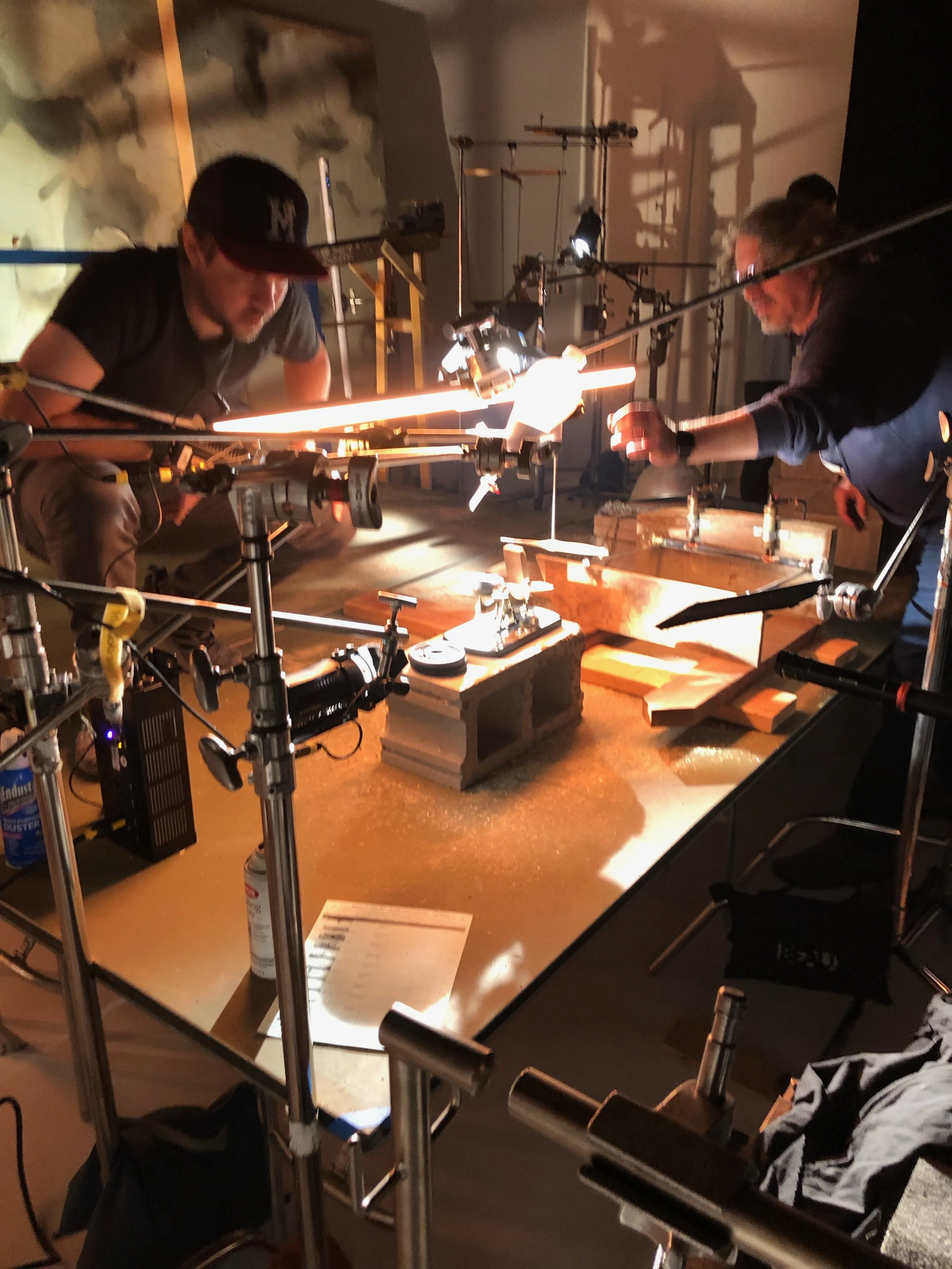 A film crew set-up with two men in a studio working under bright lights. The studio has tools, equipment, and a wooden table, with a background featuring a distressed wall.