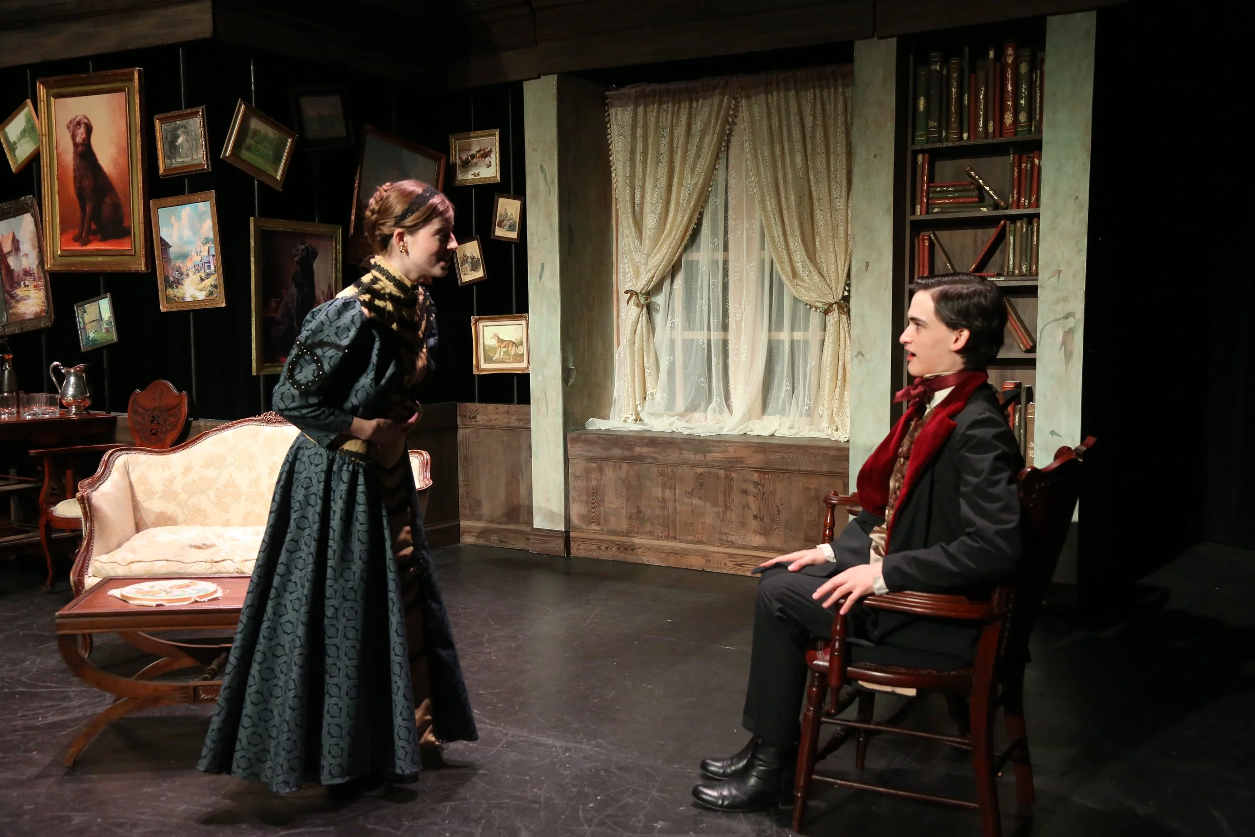 A woman with braided hair and a patterned dress talks to a seated man in a black suit with a red scarf in a vintage-style room with framed paintings on dark walls, a lace curtain window, and a bookshelf.