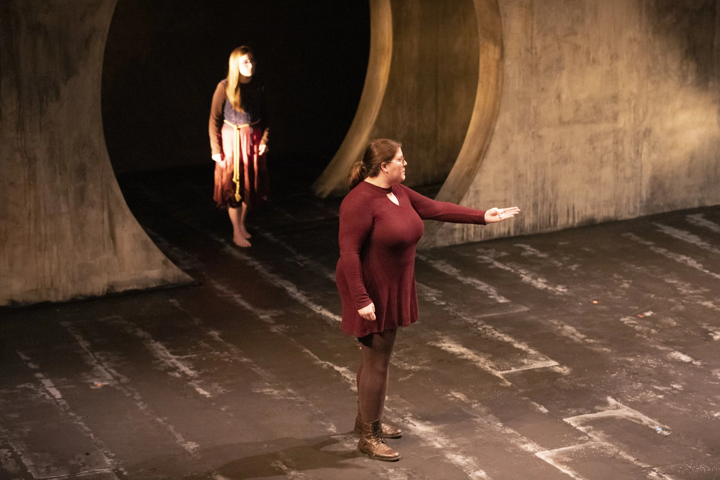 Two women in a theatrical setting; one woman in the foreground wearing a long red dress and black tights, standing with her arm extended, and another woman in the background dressed in a colorful costume, standing barefoot in a dark space framed by l
