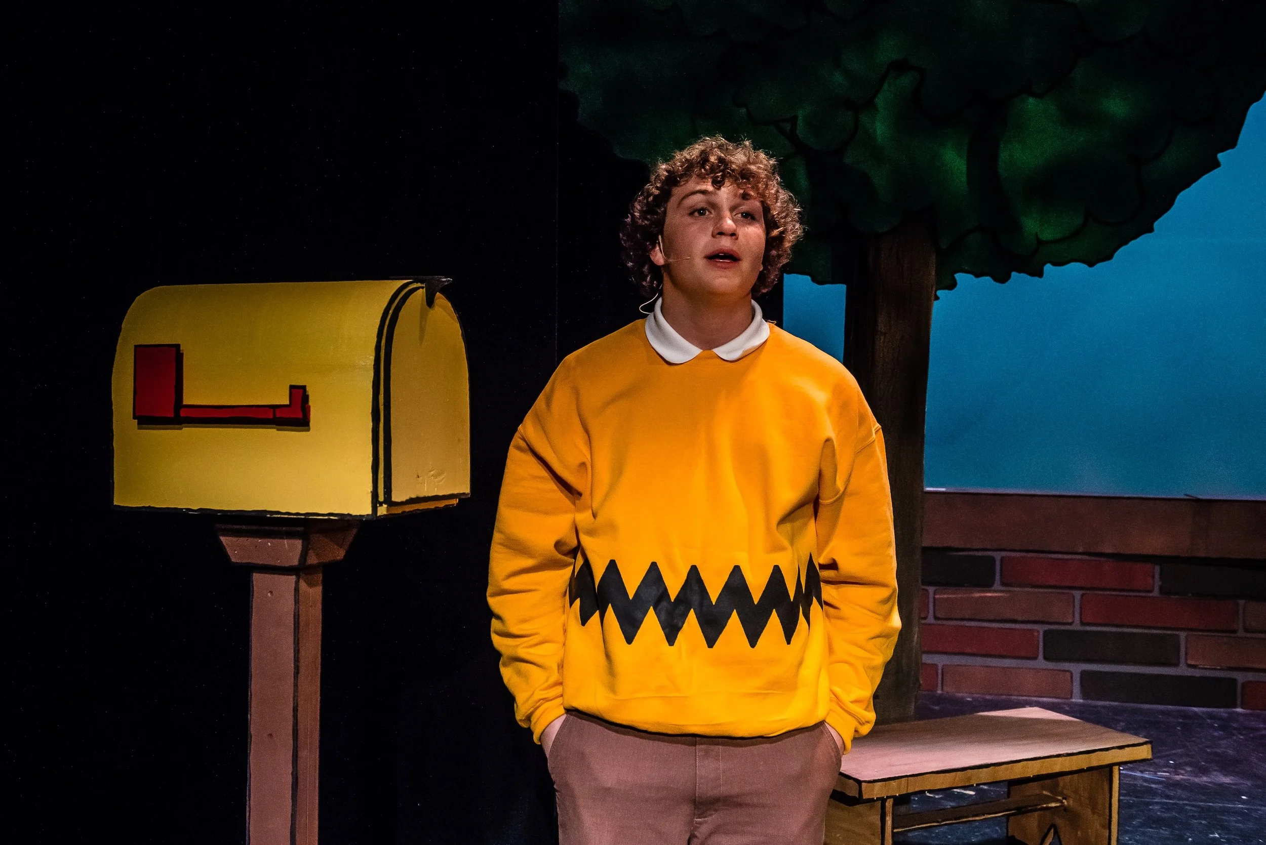 An actor on stage in a yellow costume with a black zig-zag design, standing in front of a painted backdrop of trees and a yellow mailbox.