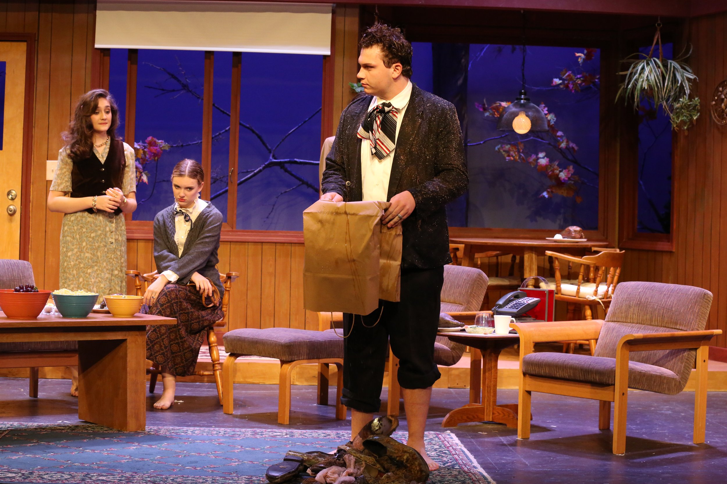 A theatrical scene with three actors on a set resembling a cozy living room with wooden paneling and large windows. Performers are sitting or standing around a table with bowls of food, and one man stands in front, holding a package.