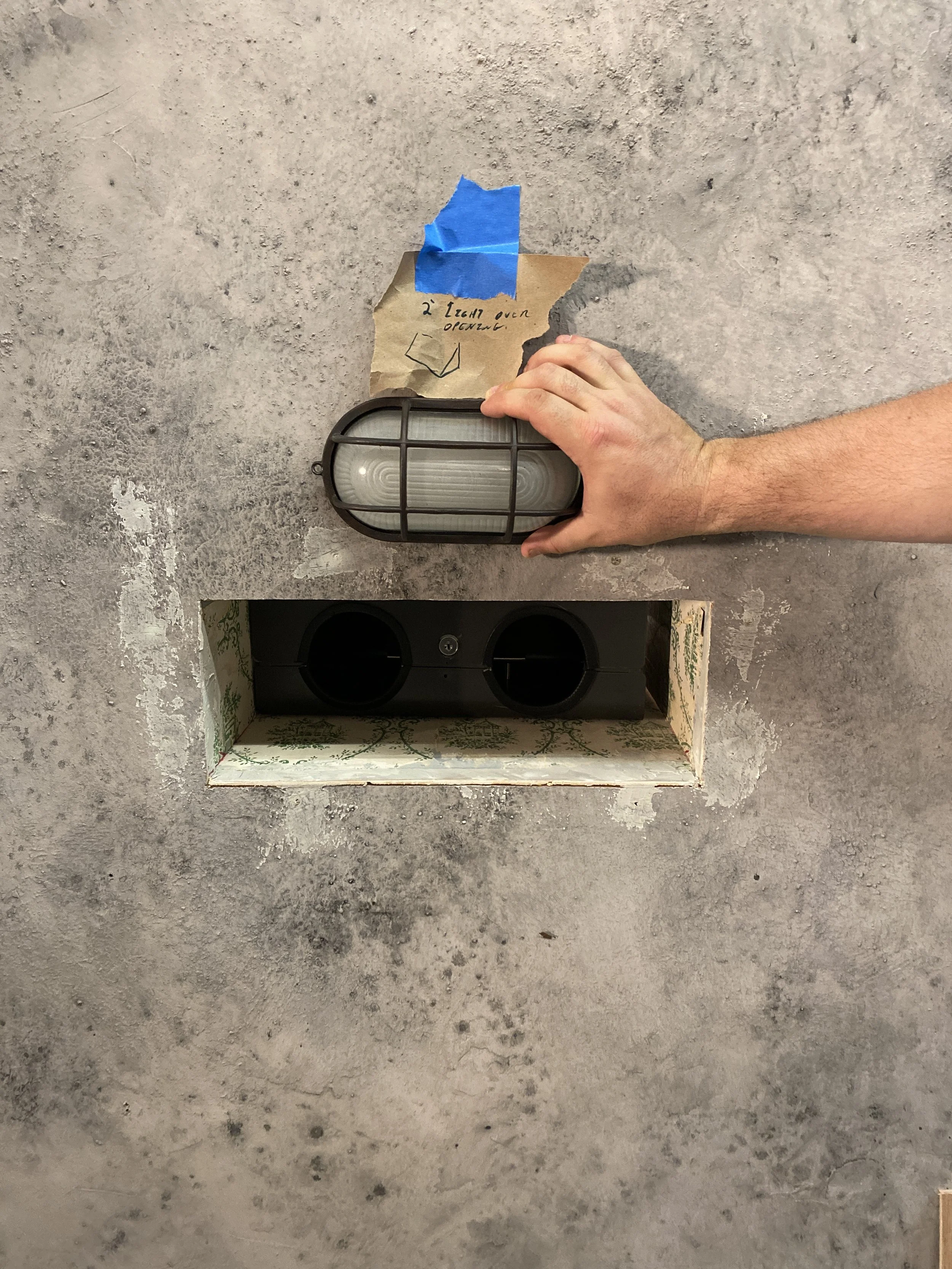 A film set with person holding a black outdoor wall light fixture on a concrete wall. There is a piece of crumpled brown paper and blue painter's tape attached to the wall near the fixture, with handwritten notes about opening the fixture.