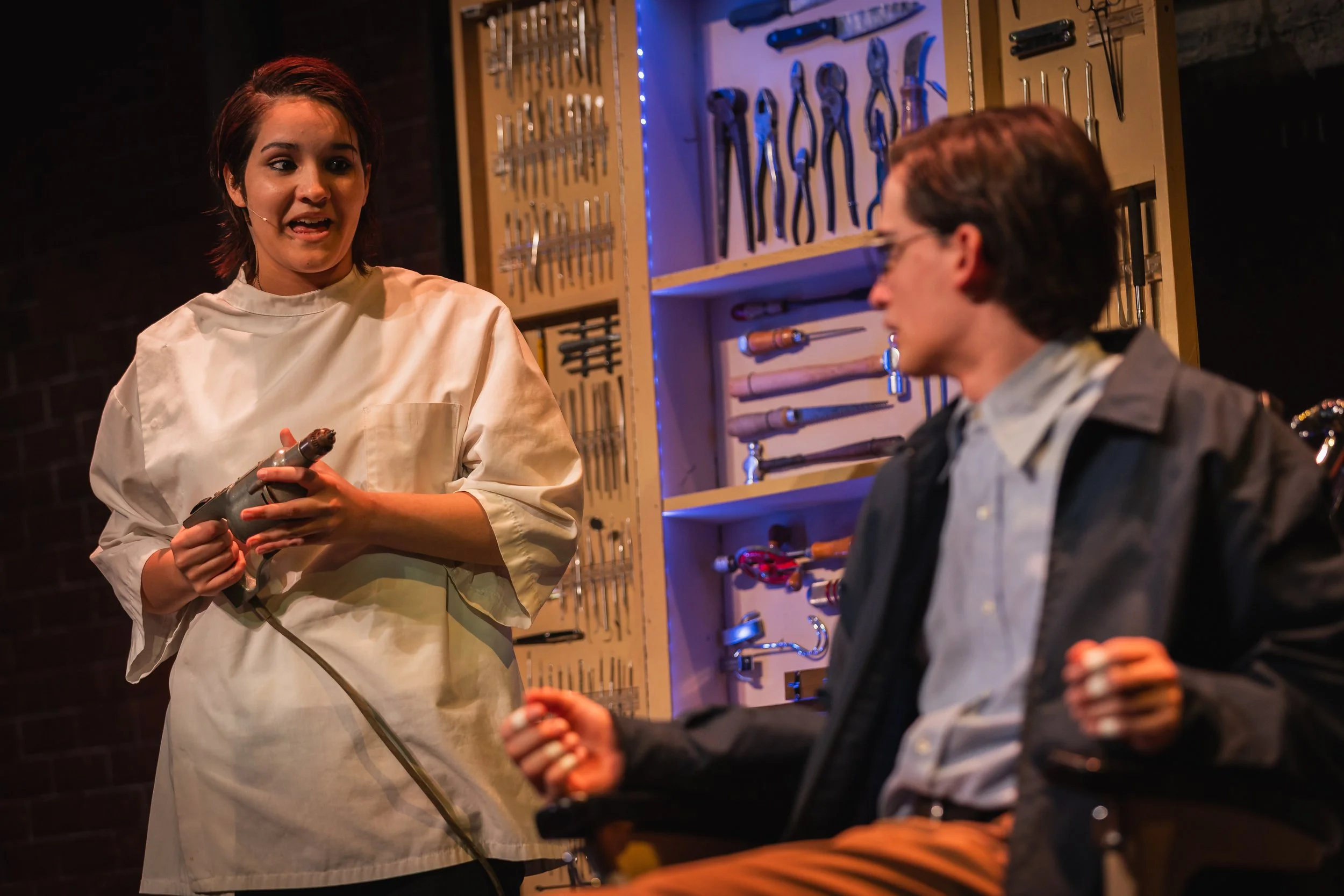 An actor portraying a dentist in a white coat holding a drill, speaking to a young man with glasses and a dark jacket, inside a set representing a dentist's office with tools on a wall in the background.