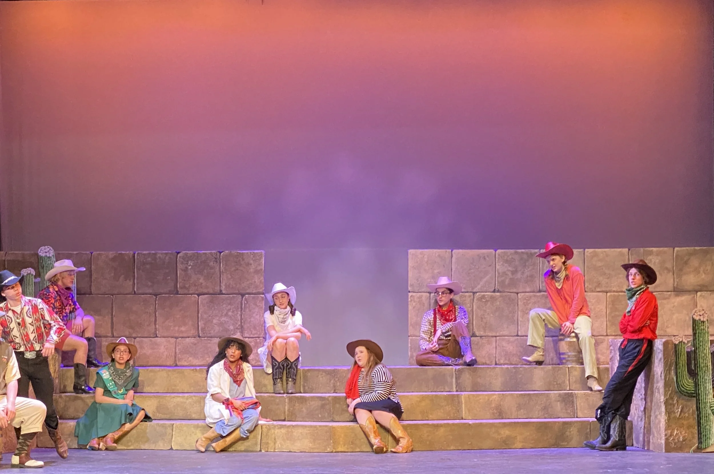 A group of actors in Western costumes performing on stage with a backdrop resembling a desert scene, including cacti and stone walls.