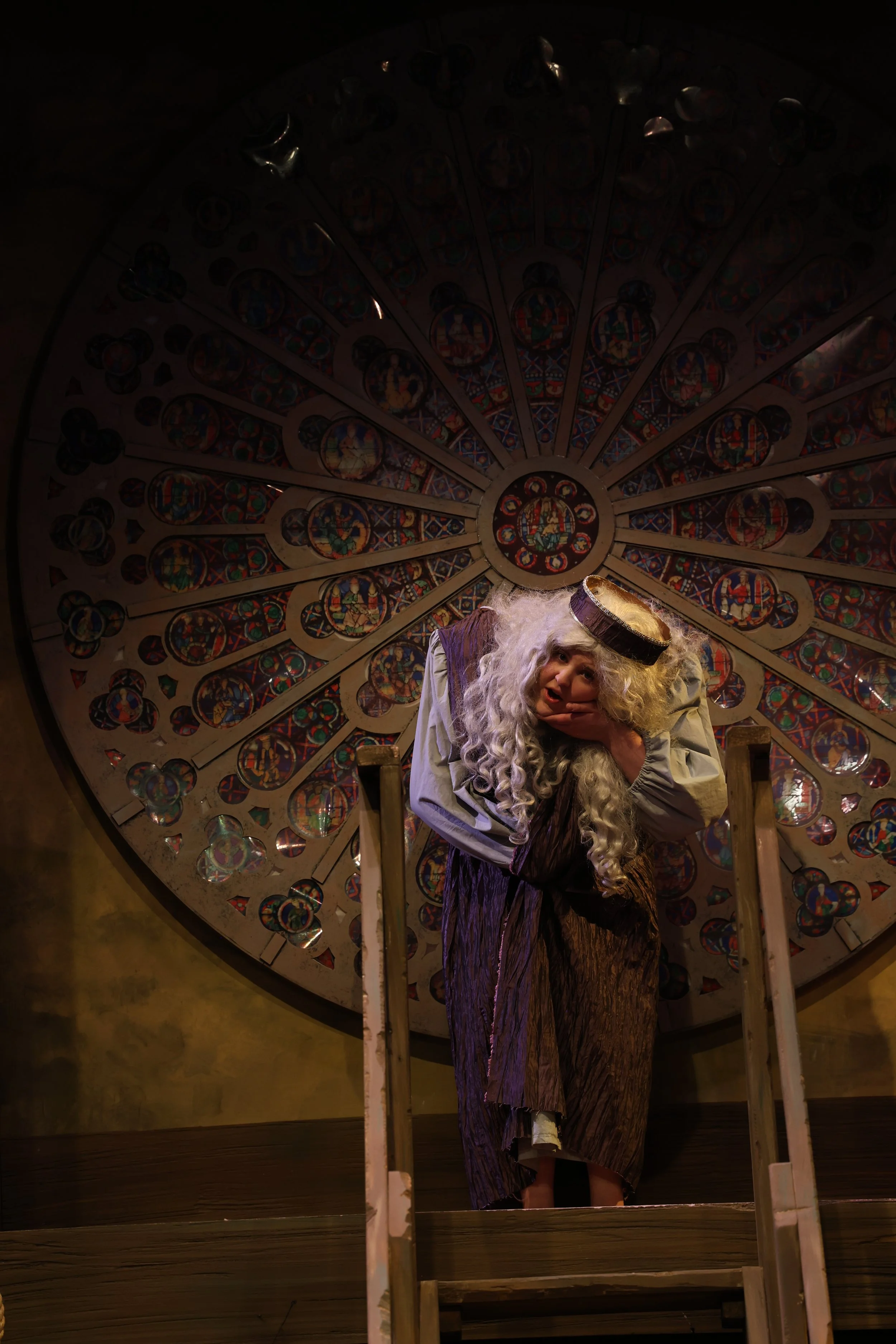 A person dressed as a saint with a long, curly white beard and a gold halo, standing on a small stage in front of a large, ornate circular stained glass window and holding their face in one hand.