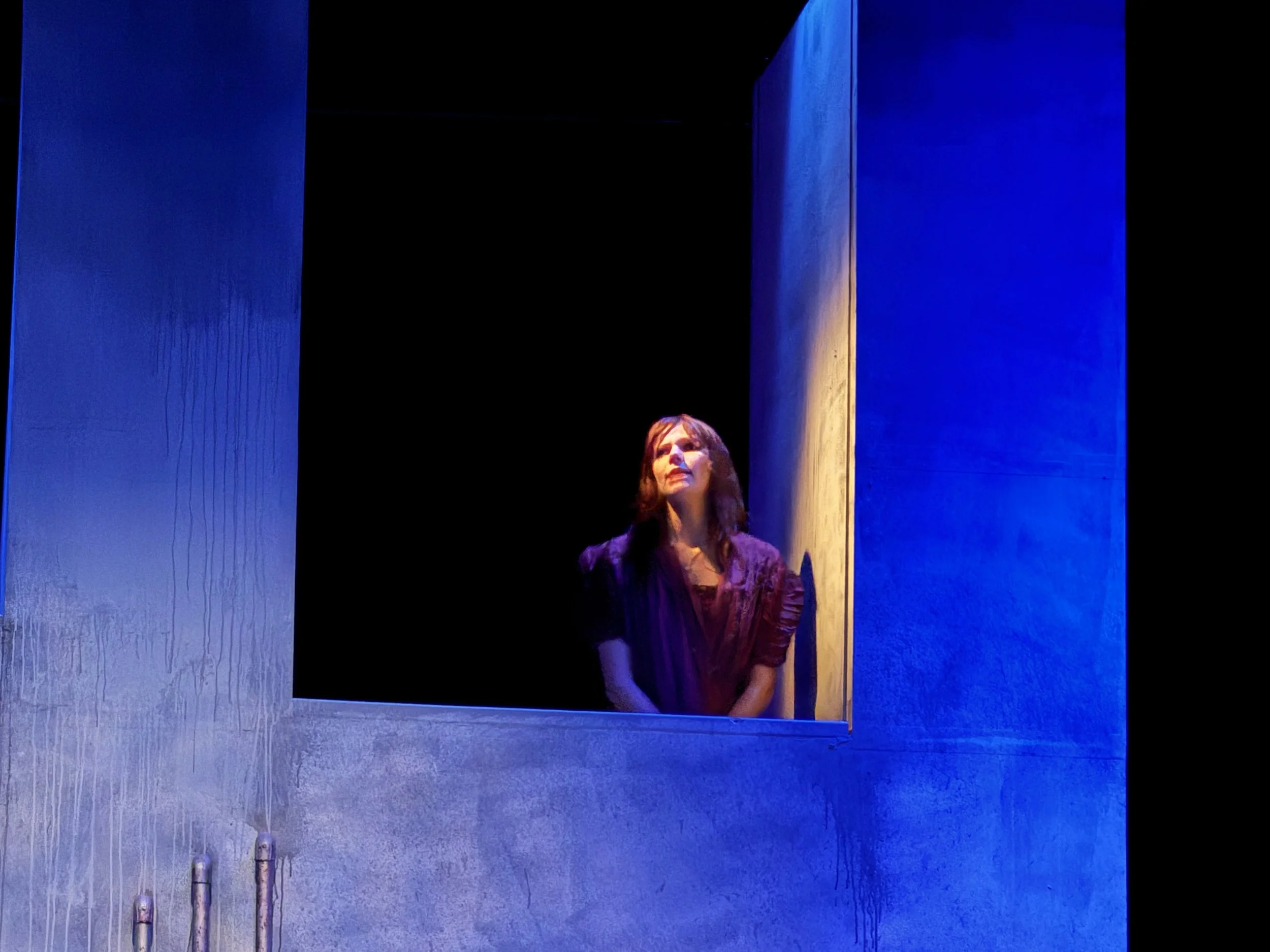 A woman with brown hair wearing a dark top stands inside a dark space, framed by a large blue and tan concrete structure, with blue lighting illuminating the scene.