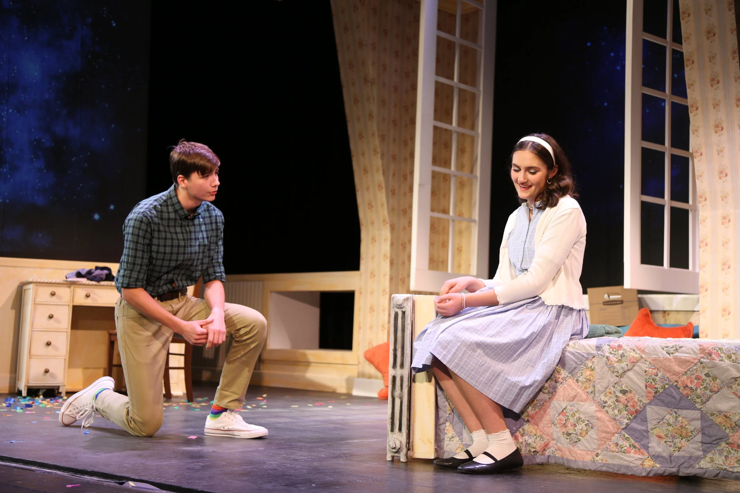 Two young actors performing on stage in a theatrical production, with a boy kneeling and a girl sitting on a bed, set against a bedroom backdrop.