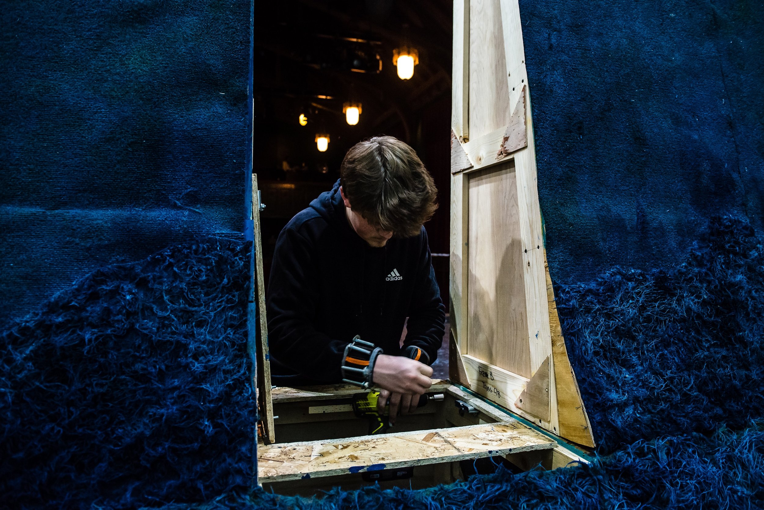 Student constructing scenery on stage, with blue textured walls and hanging lamps in the background.