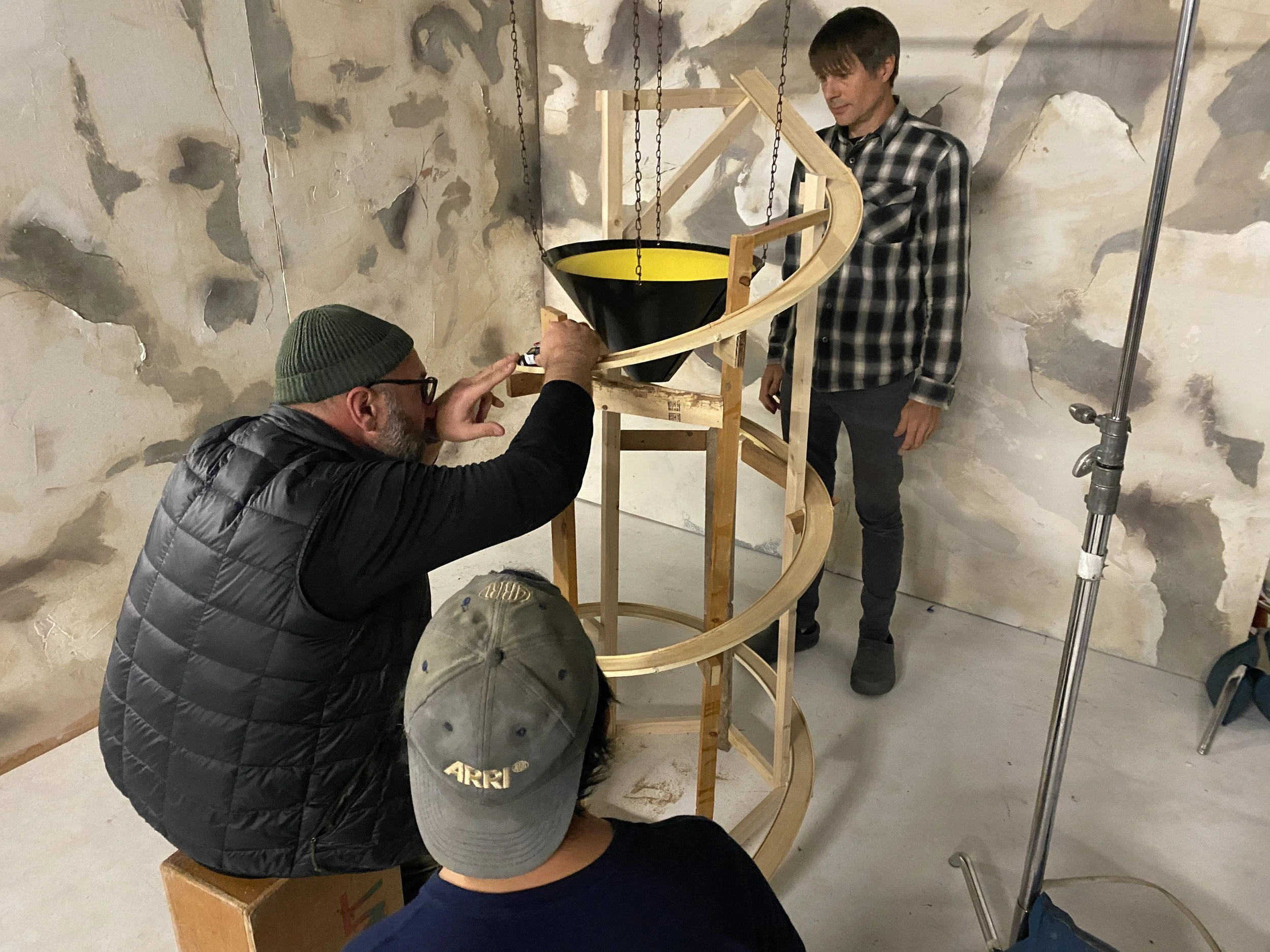 A film crew working on a wooden spiral structure in a room with peeling paint walls. One person is kneeling and adjusting something on the structure, two others are standing nearby, observing or assisting.